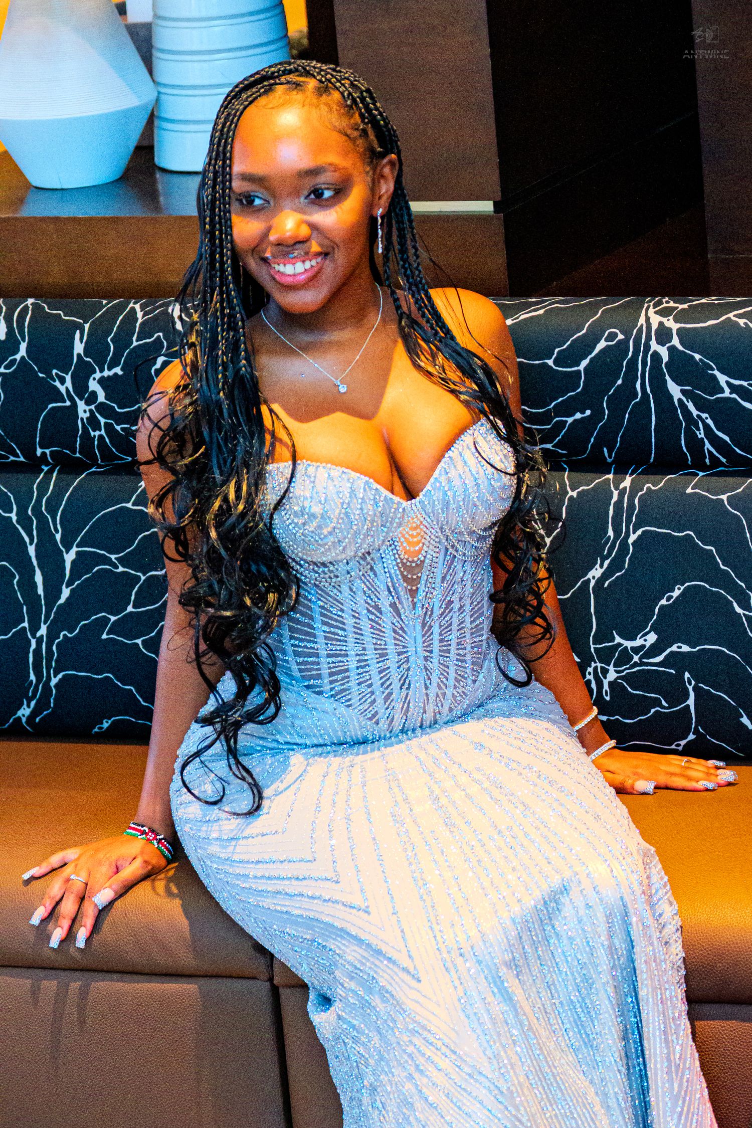 Smiling woman in beaded blue strapless gown with long braids sits on dark sofa.