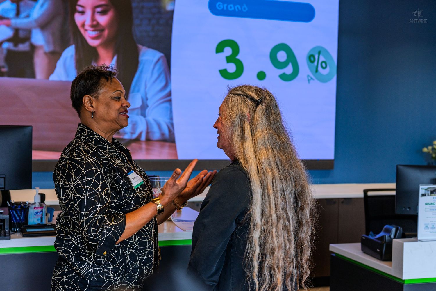 Two women conversing at a bank counter with a 3.9% interest rate displayed on a screen behind them.