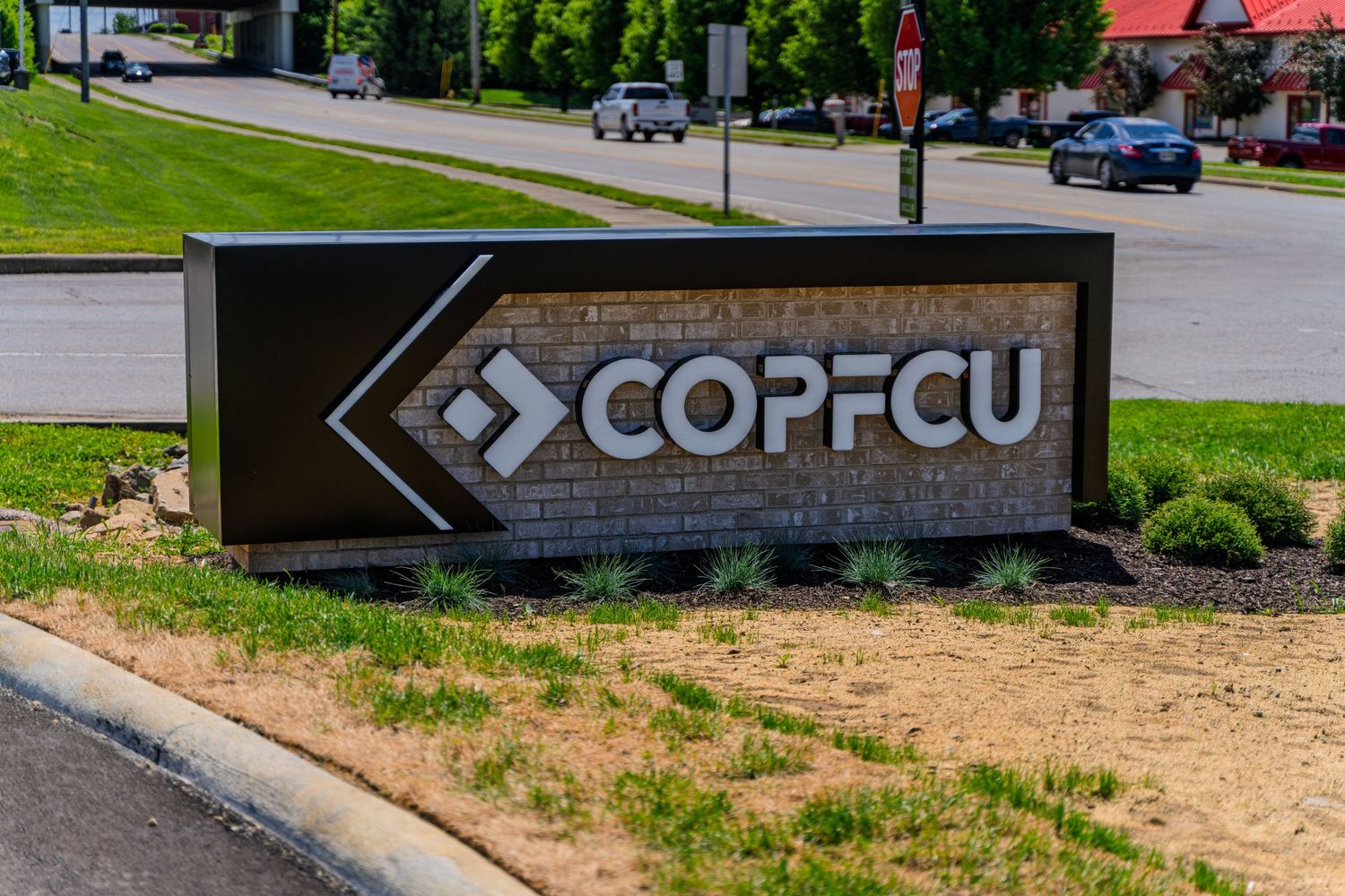 COPFCU credit union outdoor monument sign with brick and dark metal frame in a landscaped setting.