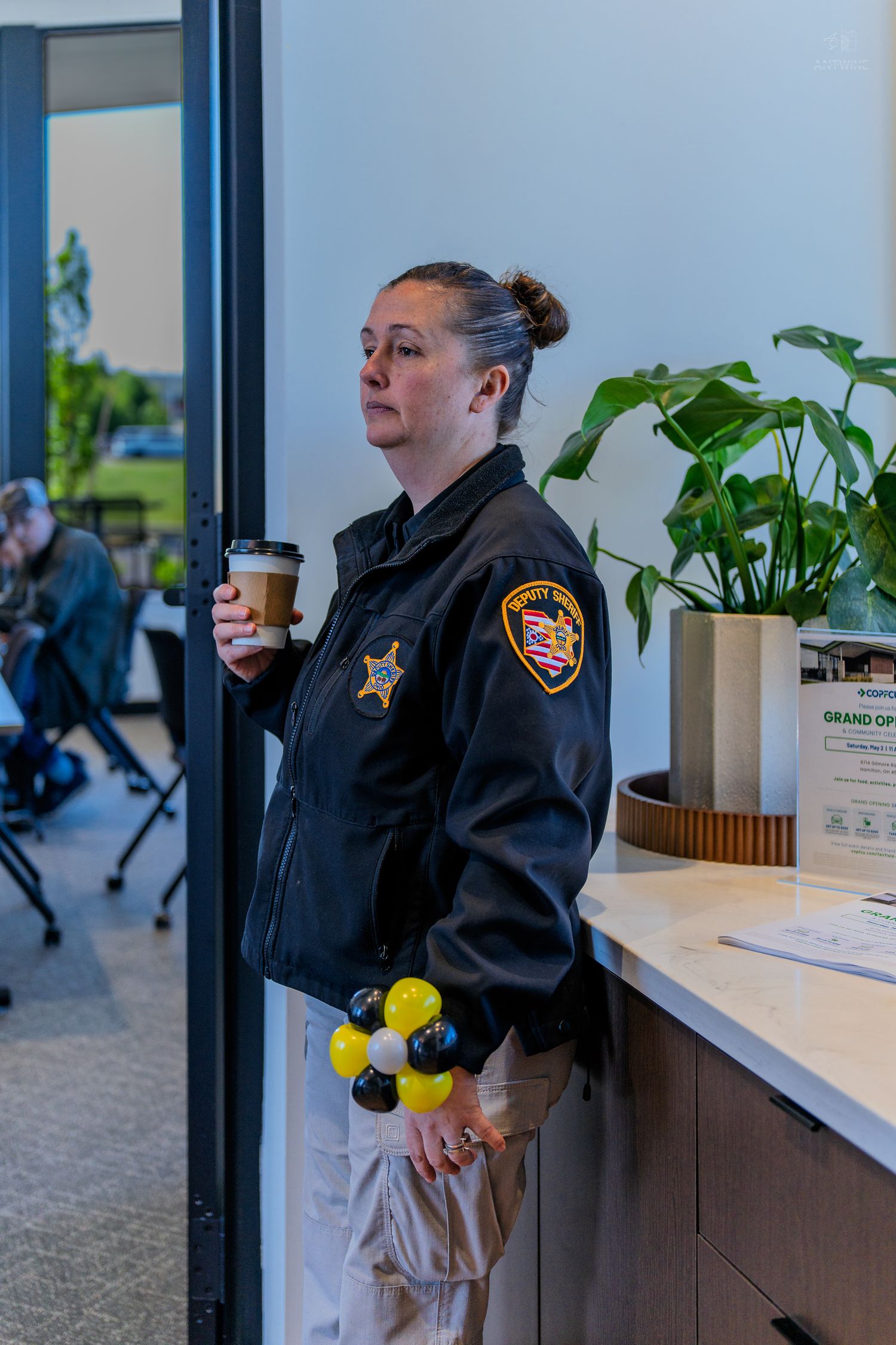 Female sheriff deputy in black uniform holds coffee cup while standing at a reception desk during a grand opening event.