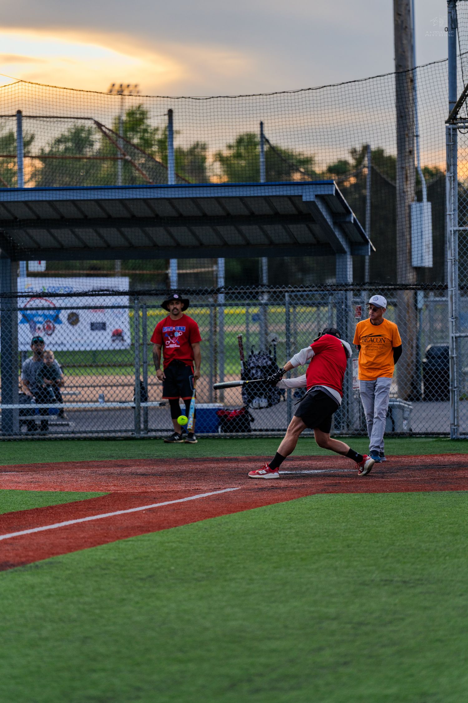 Softball batter swinging at pitch during evening recreational game at lit baseball diamond with players and dugout.