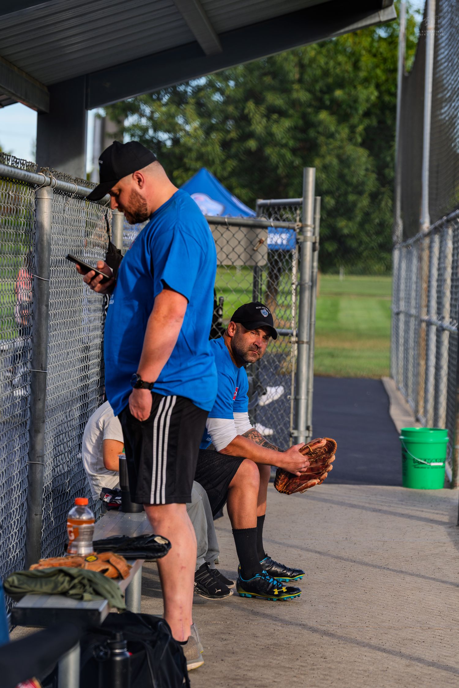 Two men in blue shirts rest in a baseball dugout, one checking his phone and one holding a glove.