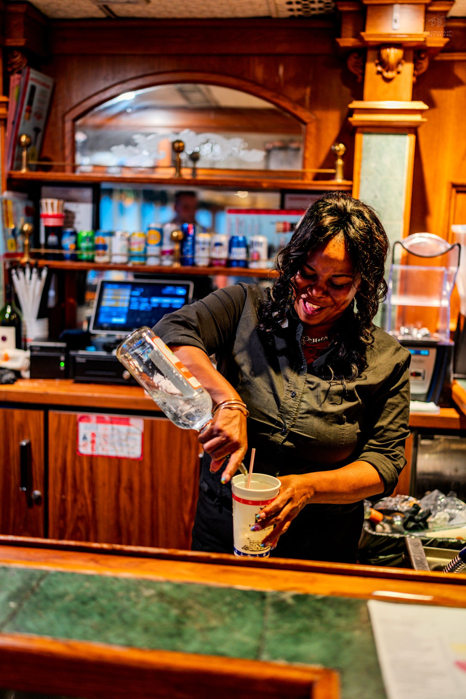 Smiling female bartender pouring a drink behind a warm wooden bar counter in a cozy restaurant setting.