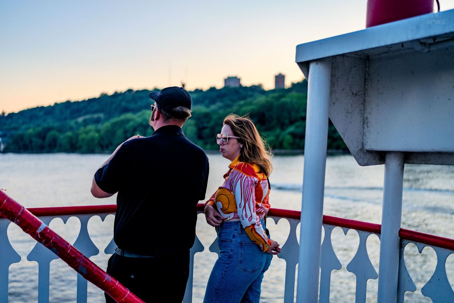 Couple stands at railing of riverboat enjoying scenic water and hillside views at golden hour sunset.