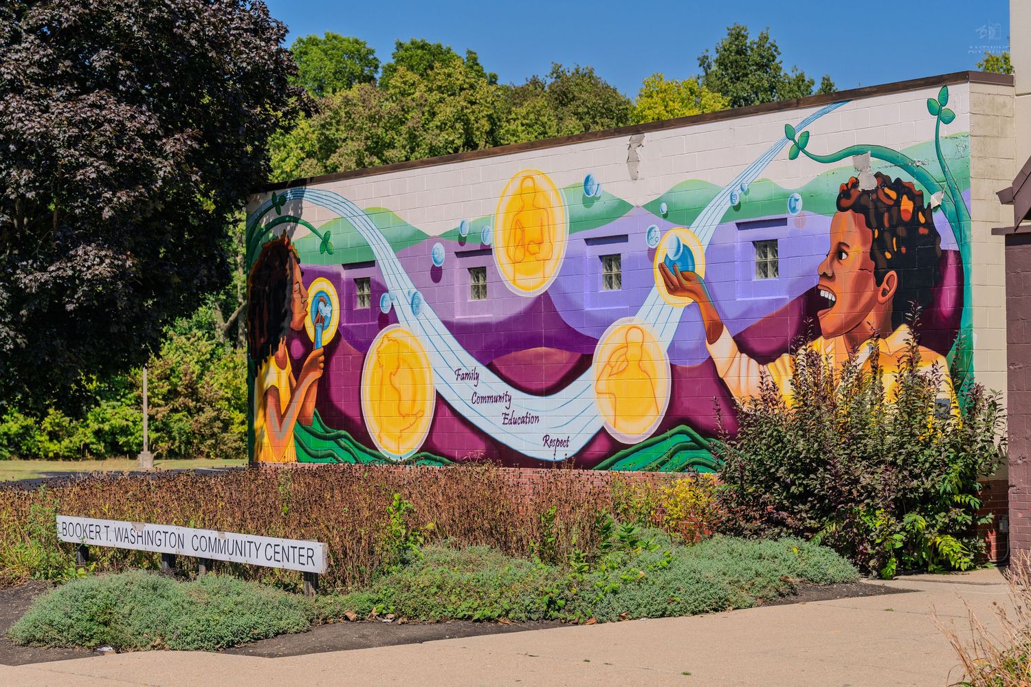 Colorful community center mural featuring vibrant figures and golden coins on an outdoor wall surrounded by greenery.