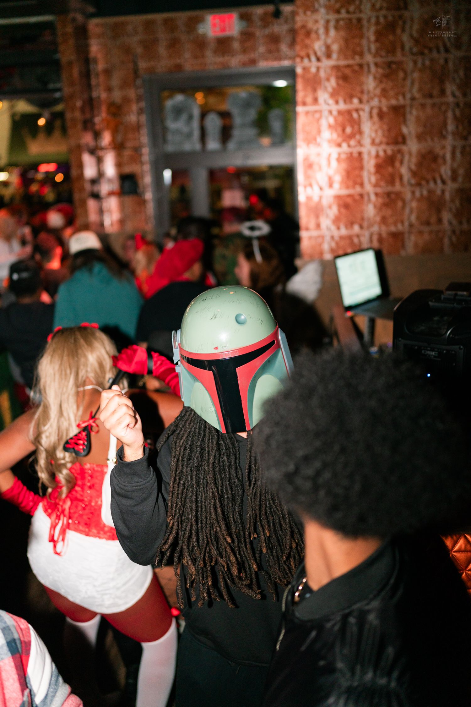 Person wearing a Boba Fett helmet costume dancing at a crowded Halloween costume party indoors.