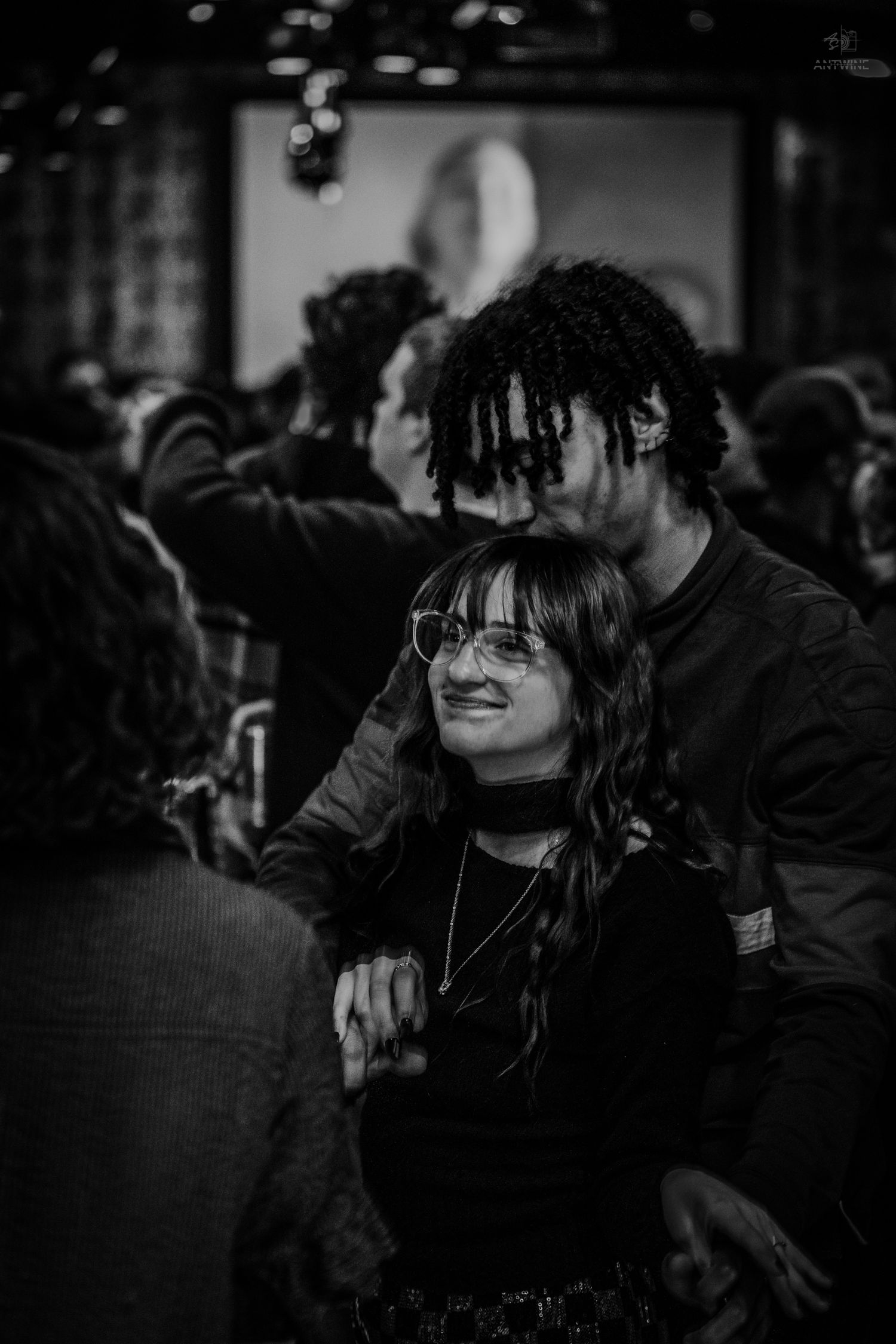 Black and white photo of a smiling woman with glasses at a crowded social event.
