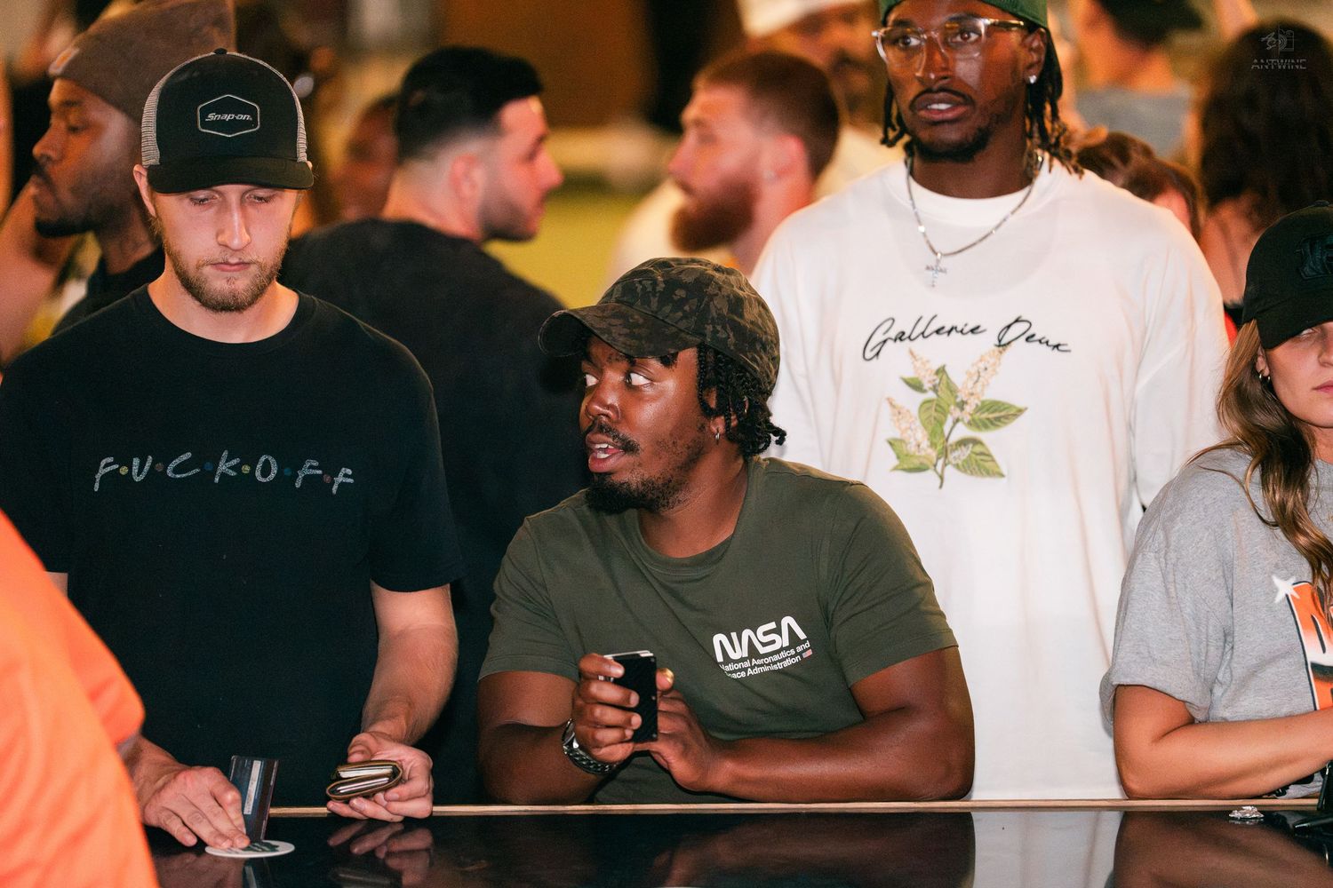 Group of men gathered around a bar counter, one wearing a NASA shirt holding a drink.