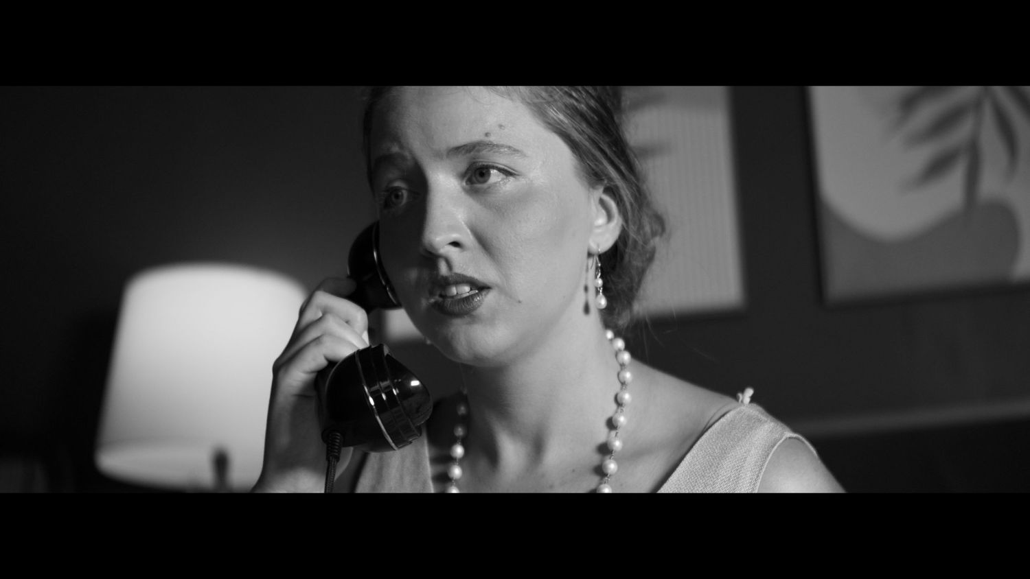 Black and white film scene of a woman in pearls speaking on a vintage telephone with dramatic lighting.