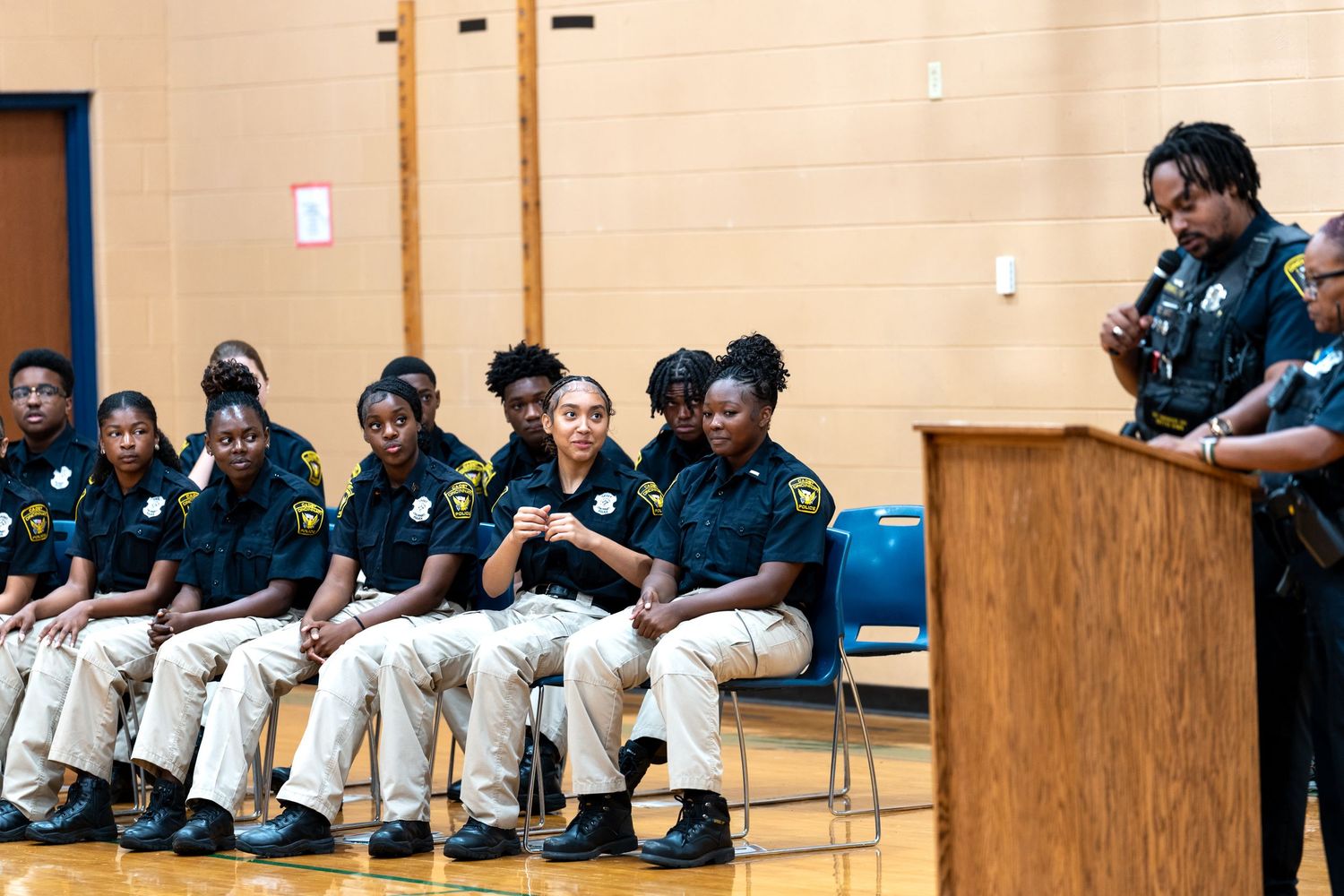 Students in matching uniforms sit in a row during a school assembly while a peer speaks at a podium.