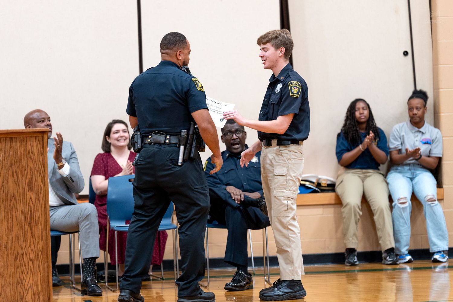 Police officer presenting certificate to young cadet at an awards ceremony while audience applauds in background.