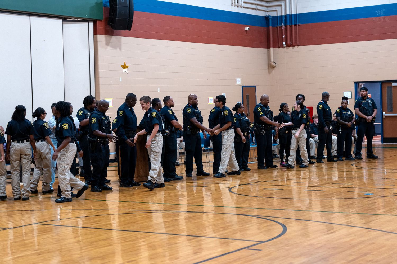 Law enforcement officers in uniform standing in a line during a ceremony inside a school gymnasium.