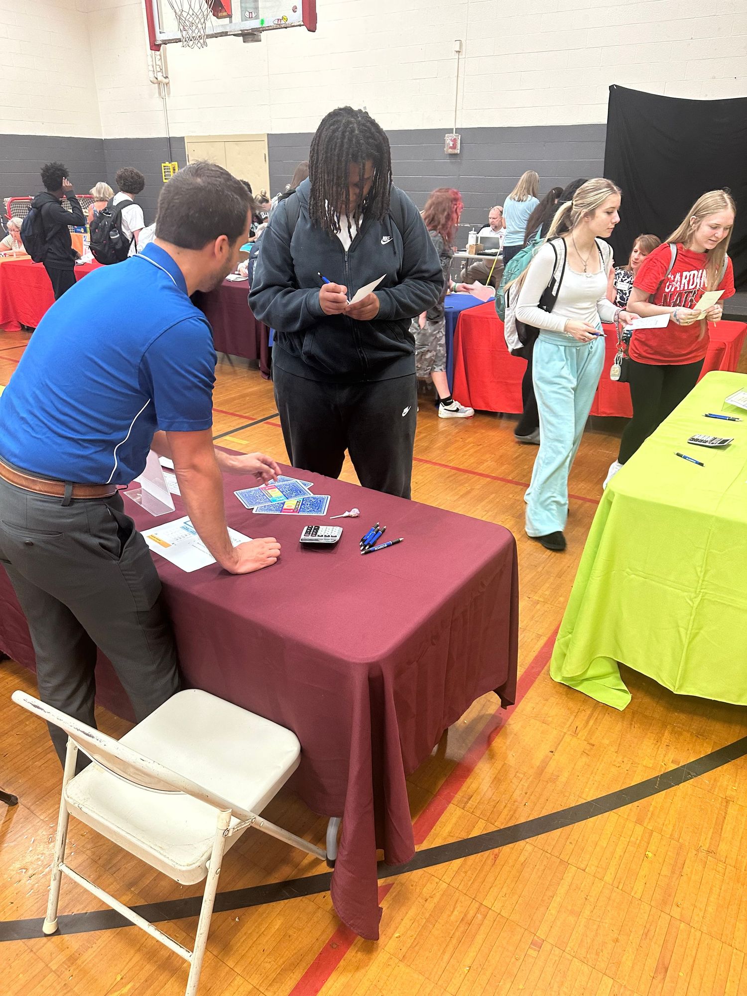 Students browse booths at an indoor school career fair with colorful tables in a gymnasium.