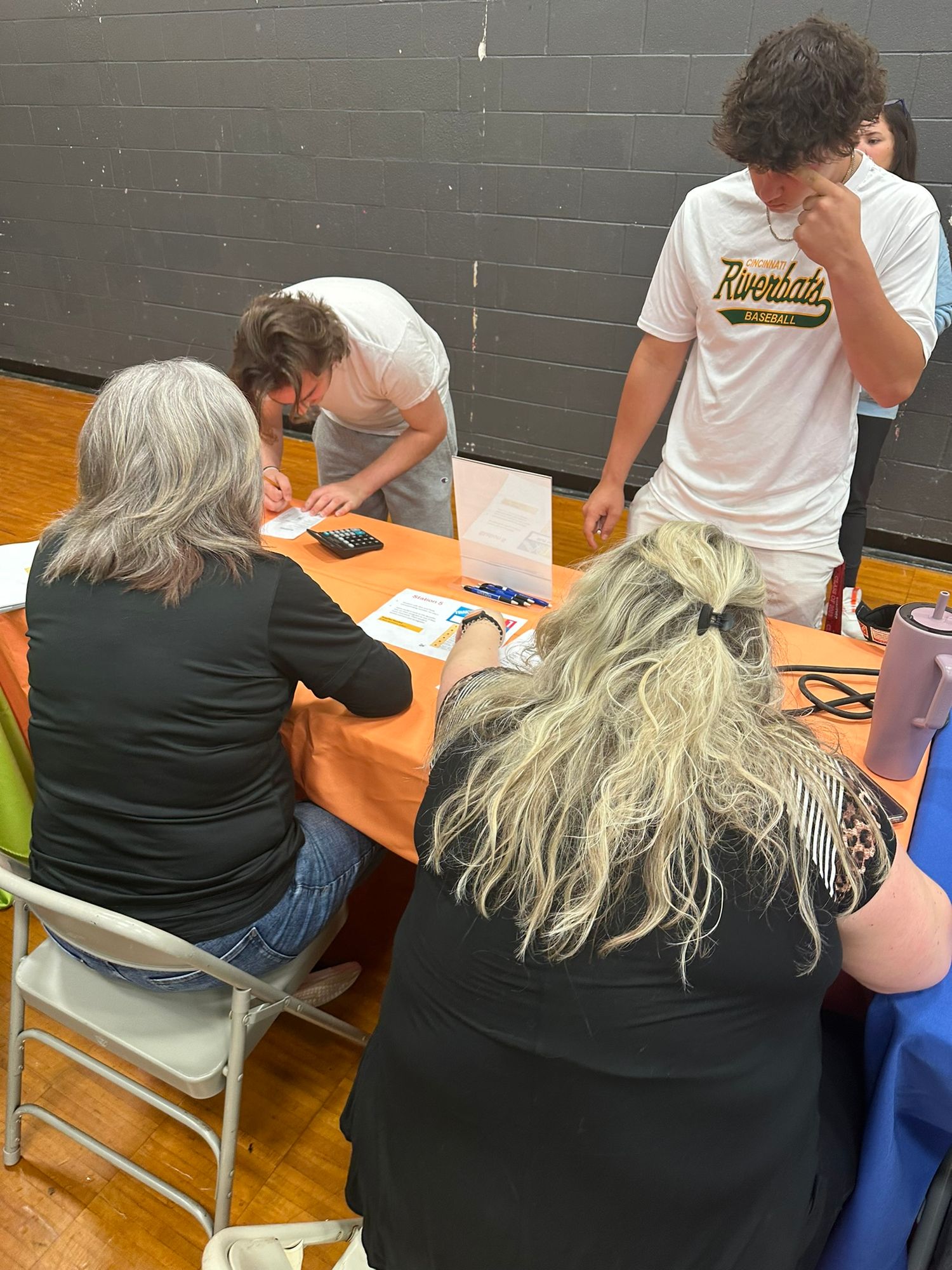 Students and adults gather around an orange table in a gym, reviewing paperwork during a school registration event.