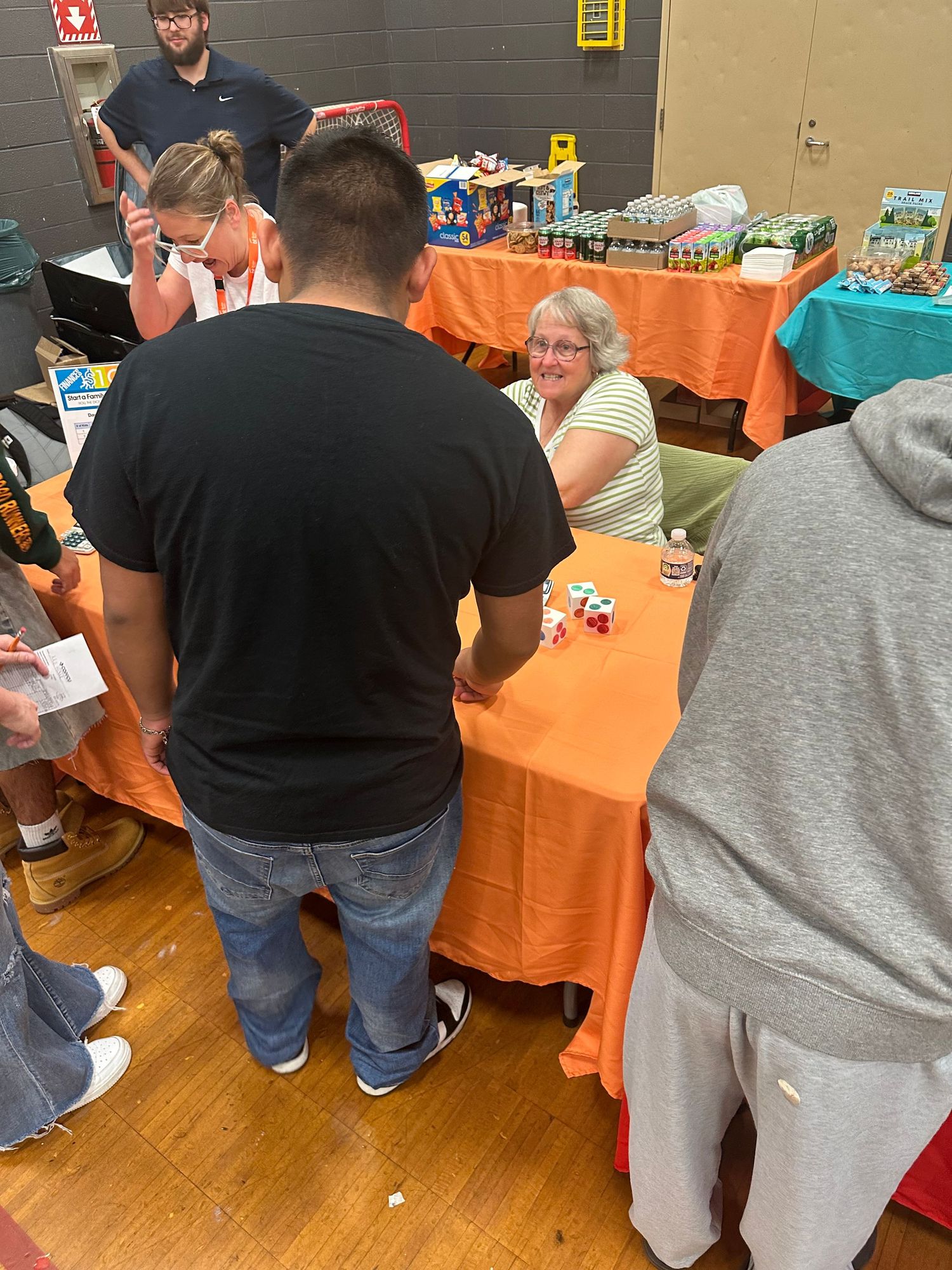 People gathered around an orange-draped table at an indoor community event, with a smiling woman seated behind it.