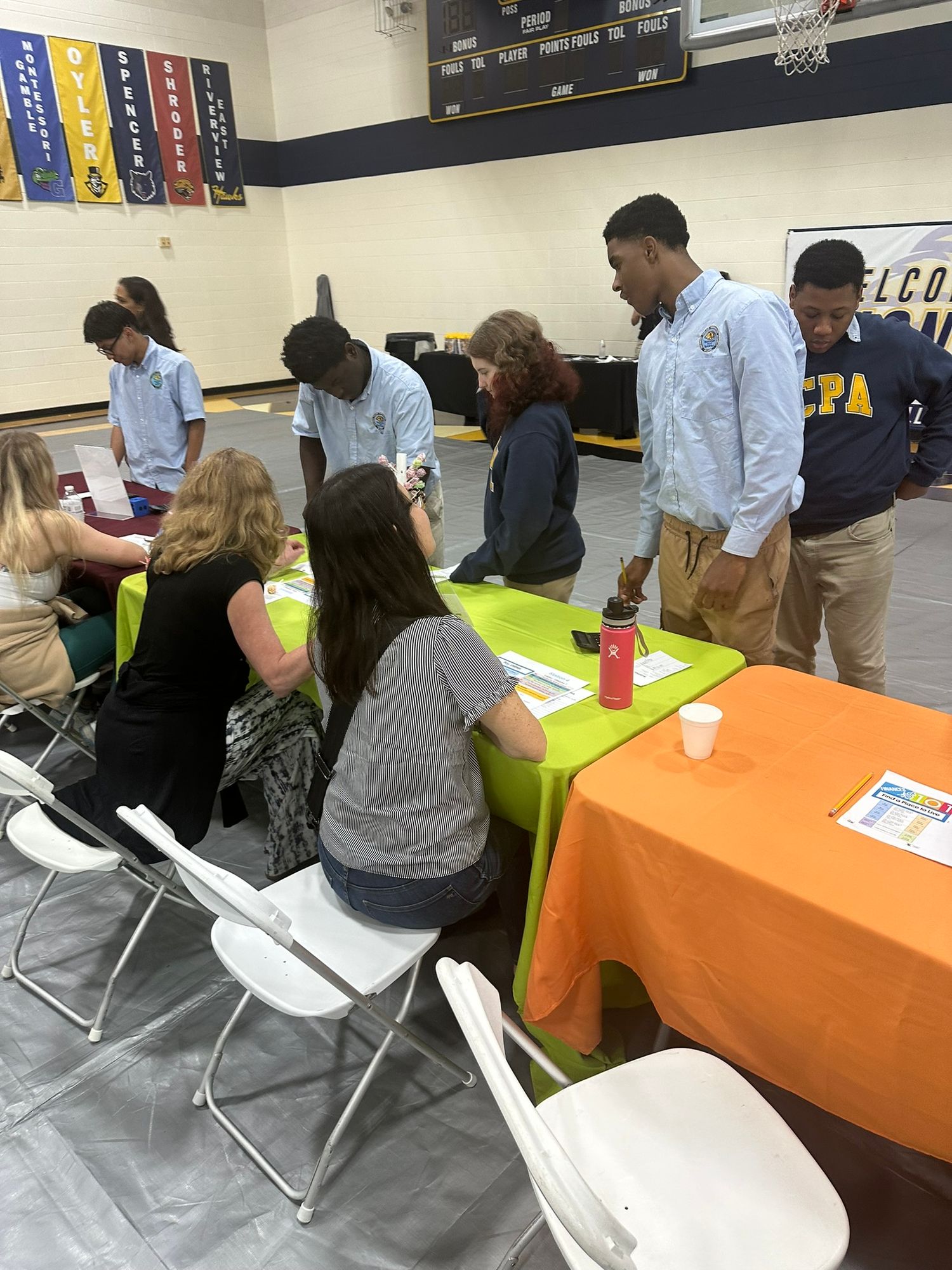 Students and teachers interact at colorful tables during a school fair event in a gymnasium decorated with pennants.