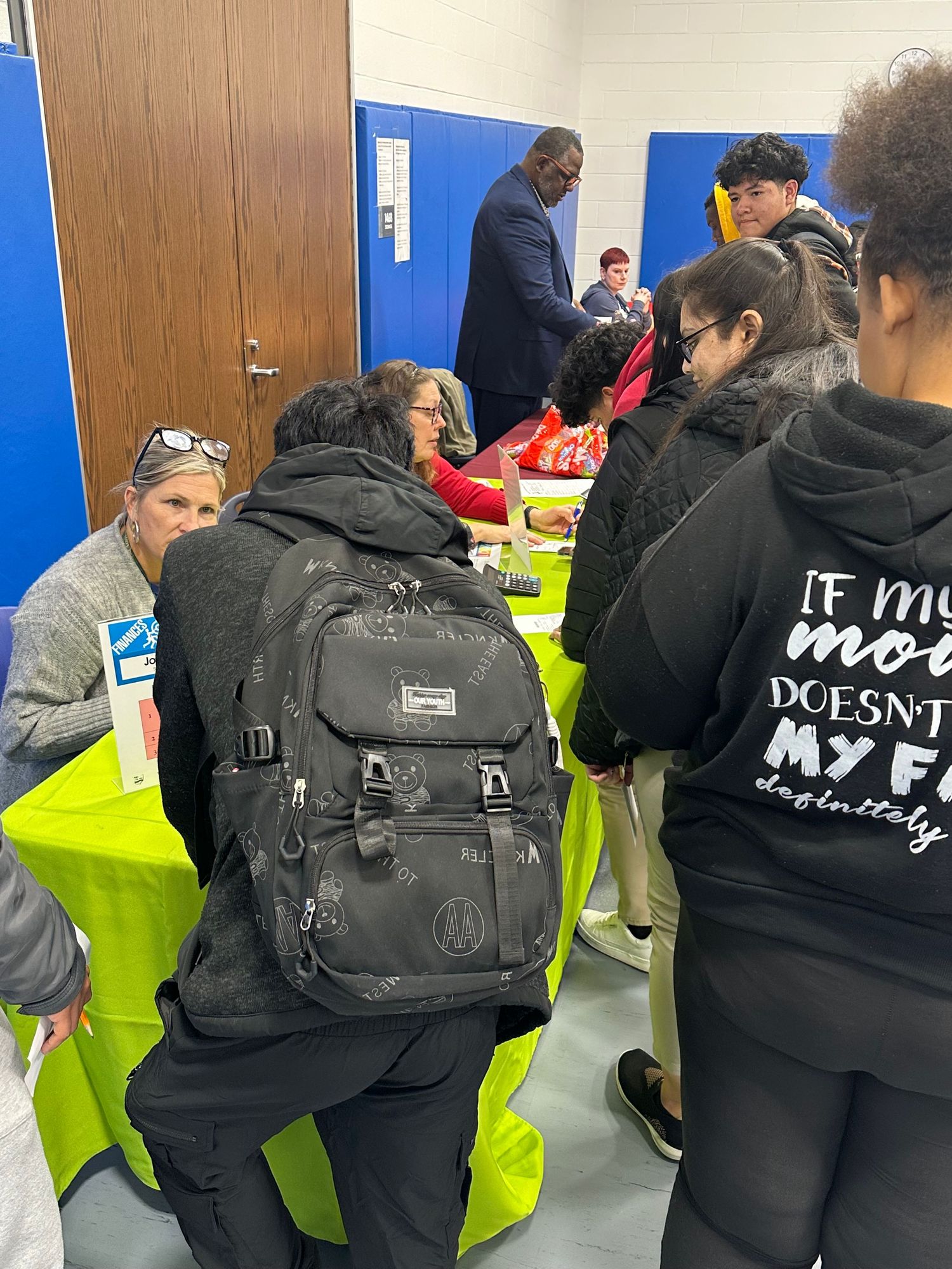 Students and adults gather around green tables at a school event, with one student wearing a black hoodie and backpack.