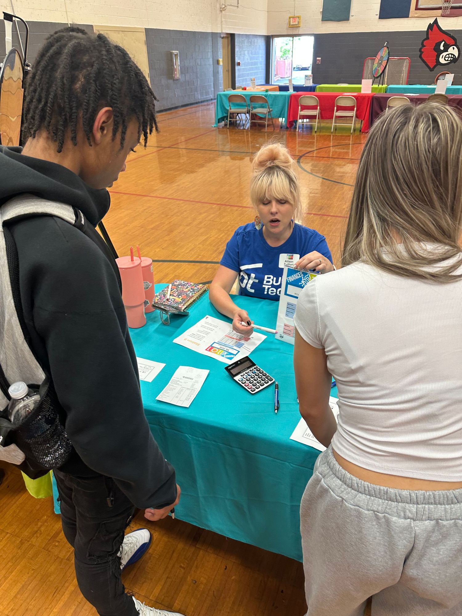 A woman in a blue shirt assists two students at a teal table during a school career fair event.