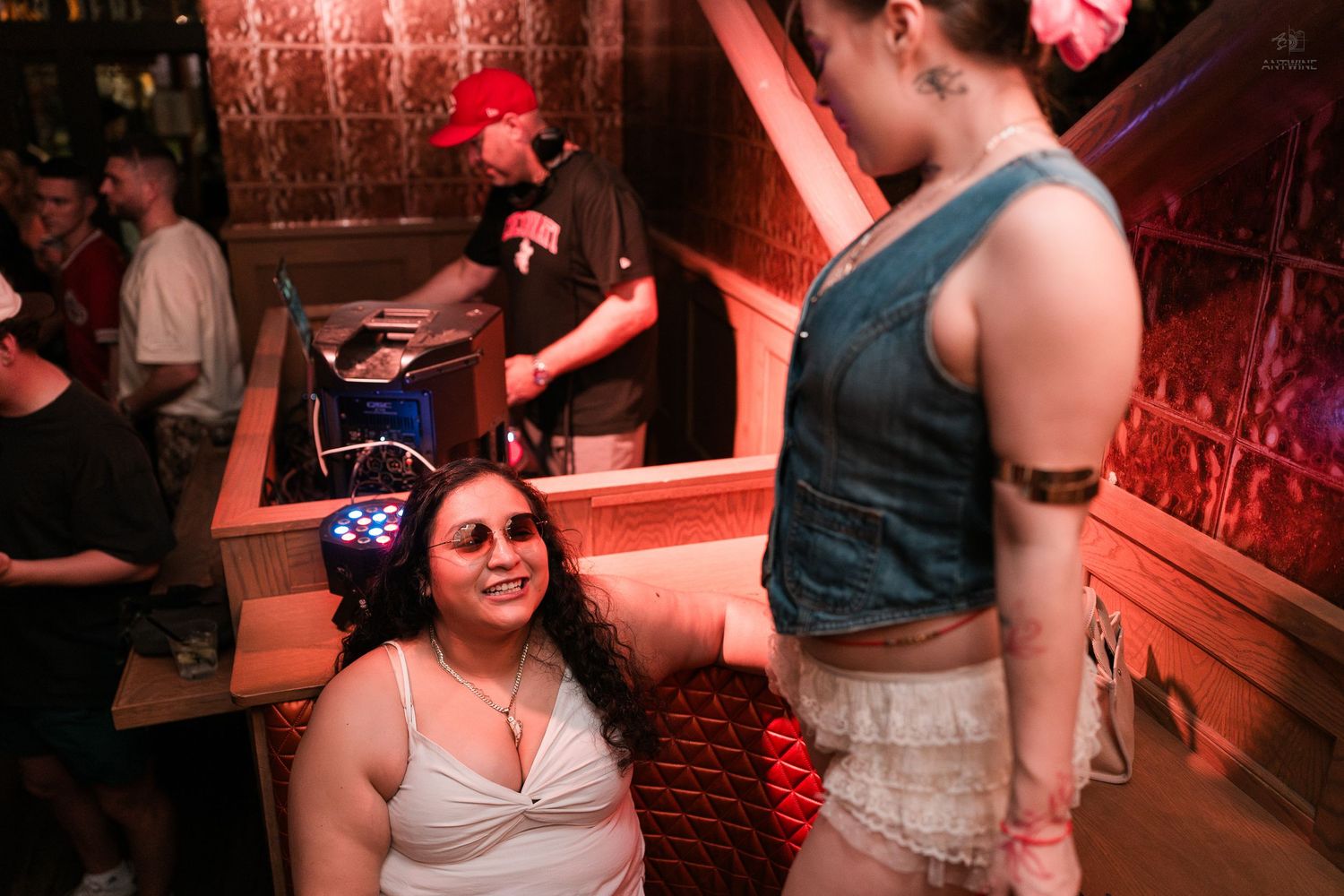 A woman wearing star-patterned glasses smiles up at another woman in a denim vest at a crowded nightclub.