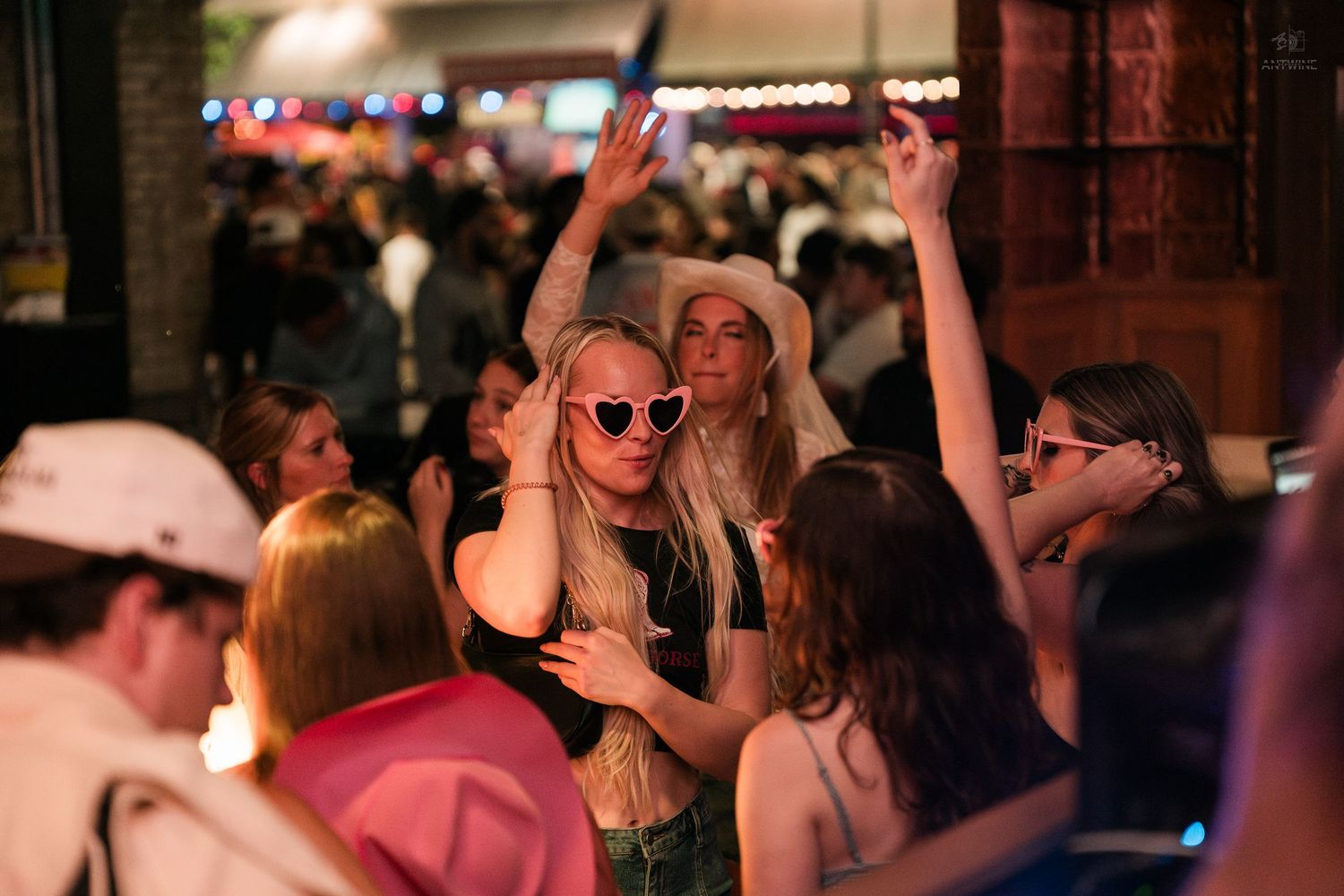 Two women dancing and celebrating at a crowded outdoor bar, one wearing heart-shaped sunglasses and a cowboy hat.