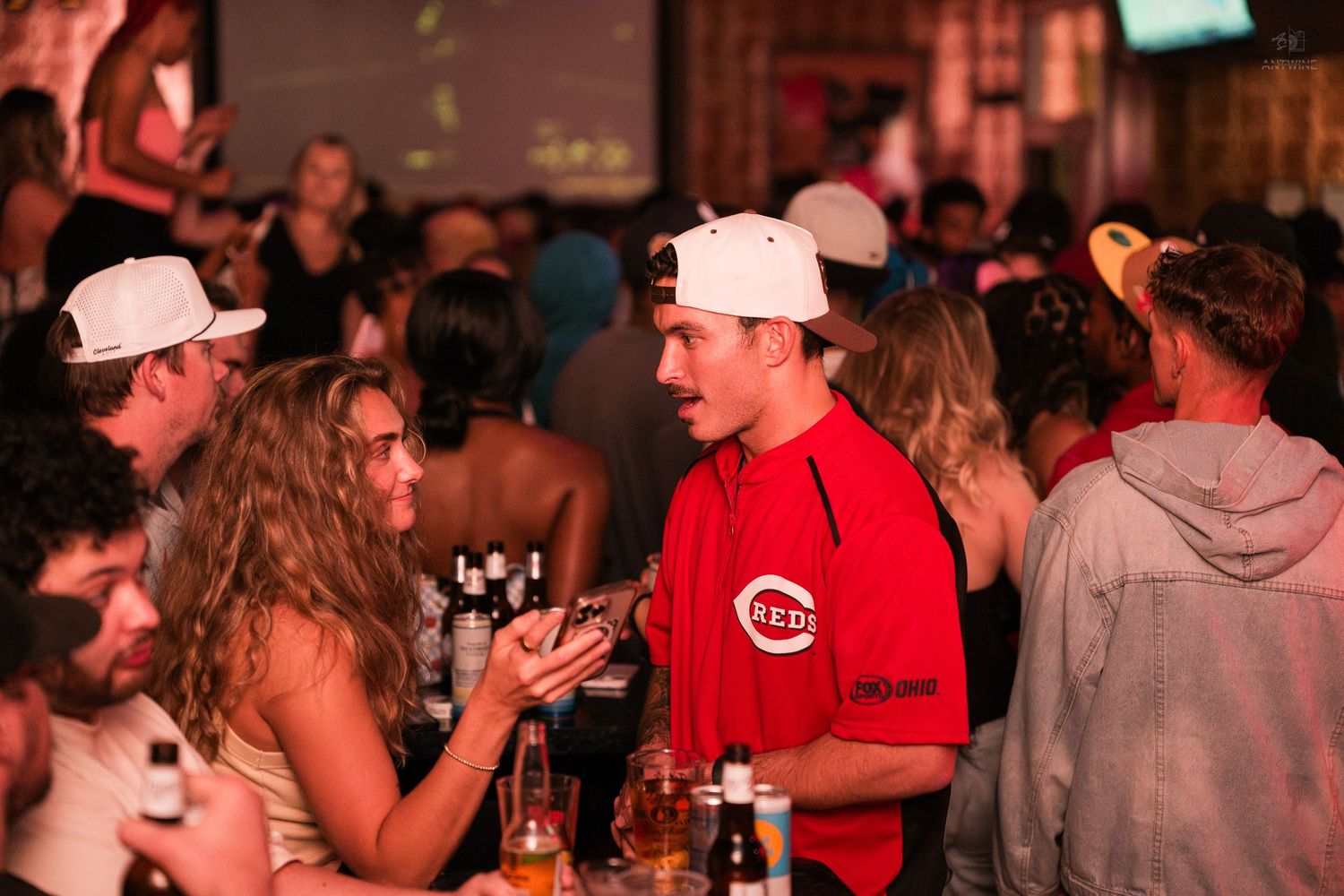 Man in Cincinnati Reds jersey and white cap socializes with woman at crowded sports bar.