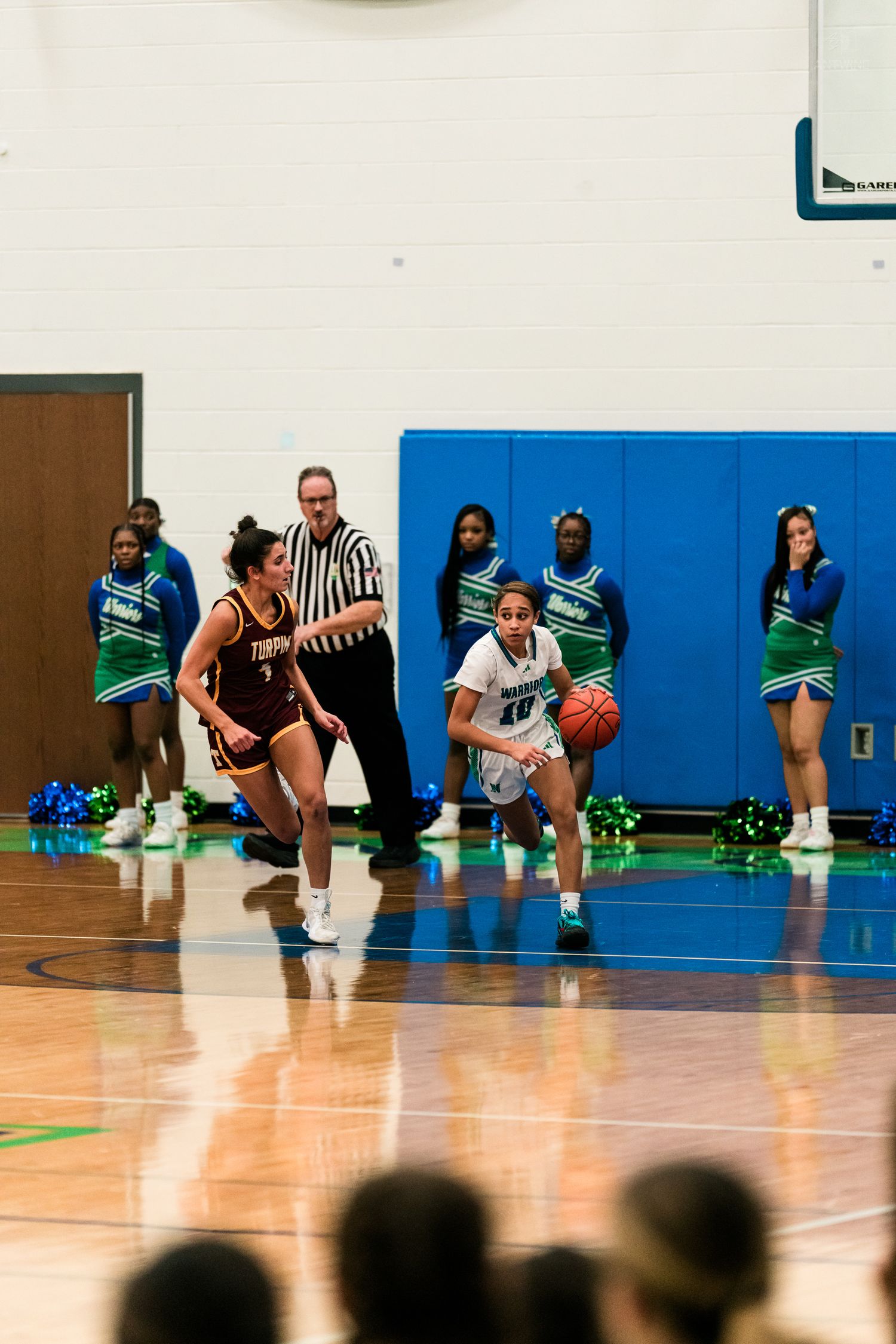 Warriors player dribbles past maroon-uniformed Trojan defender with referee and cheerleaders visible in background.