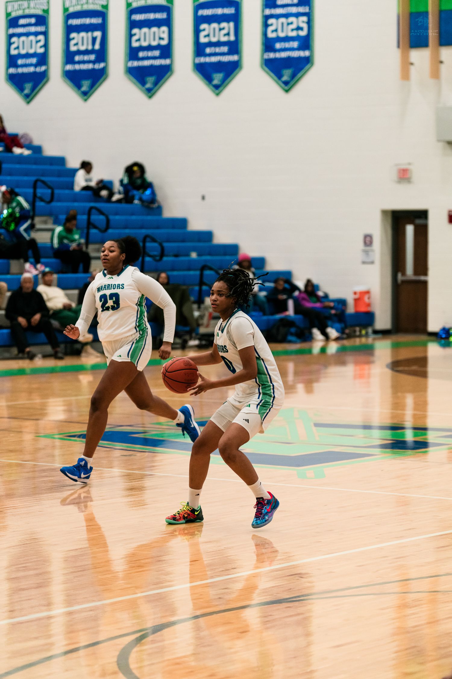 Two female basketball players in white Warriors uniforms run down court during a high school girls basketball game.