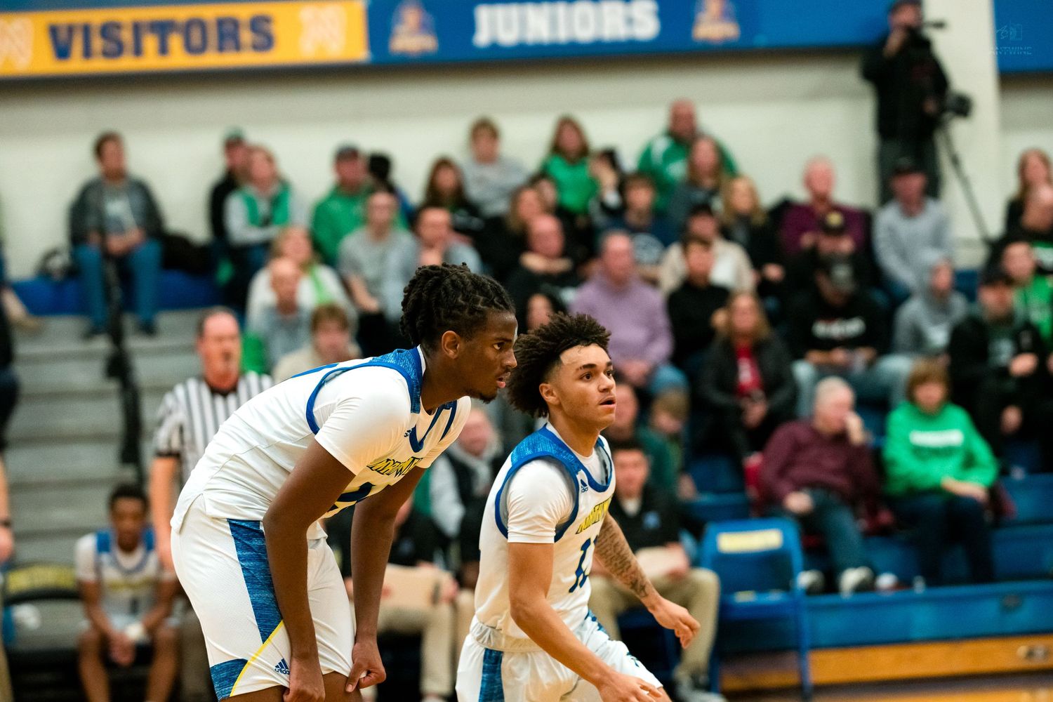 Two blue-uniformed high school basketball players set screens near the visitors section during a packed gym game.