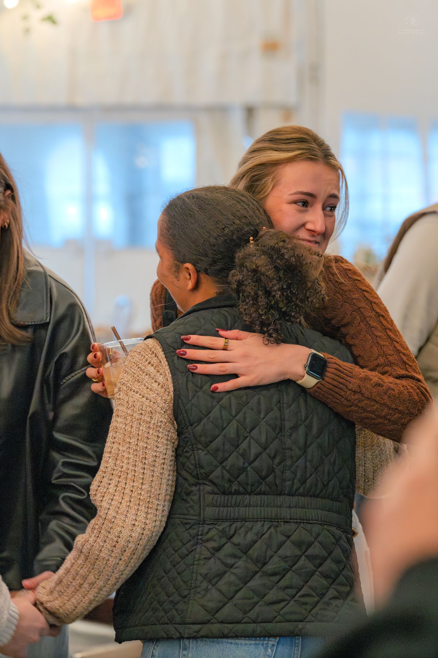 Two young women share a warm embrace at a social gathering, one smiling over the others shoulder.