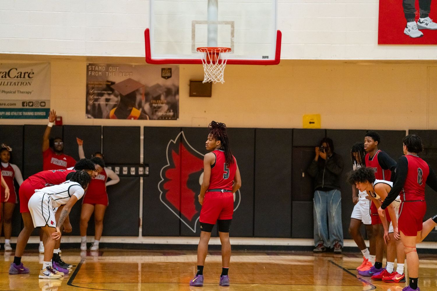 Girls basketball team practices free throws in a high school gymnasium with a cardinal mascot logo on the floor.