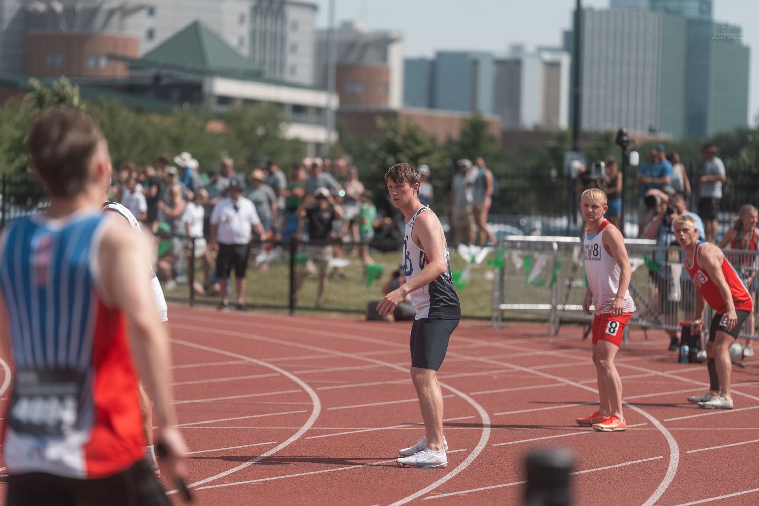 Male track athletes prepare for baton exchange during outdoor relay race on sunny day.