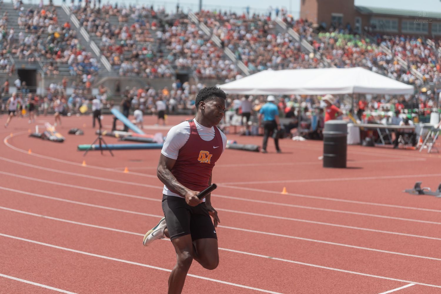 A relay runner holds a baton mid-stride at a packed outdoor track and field championship meet.
