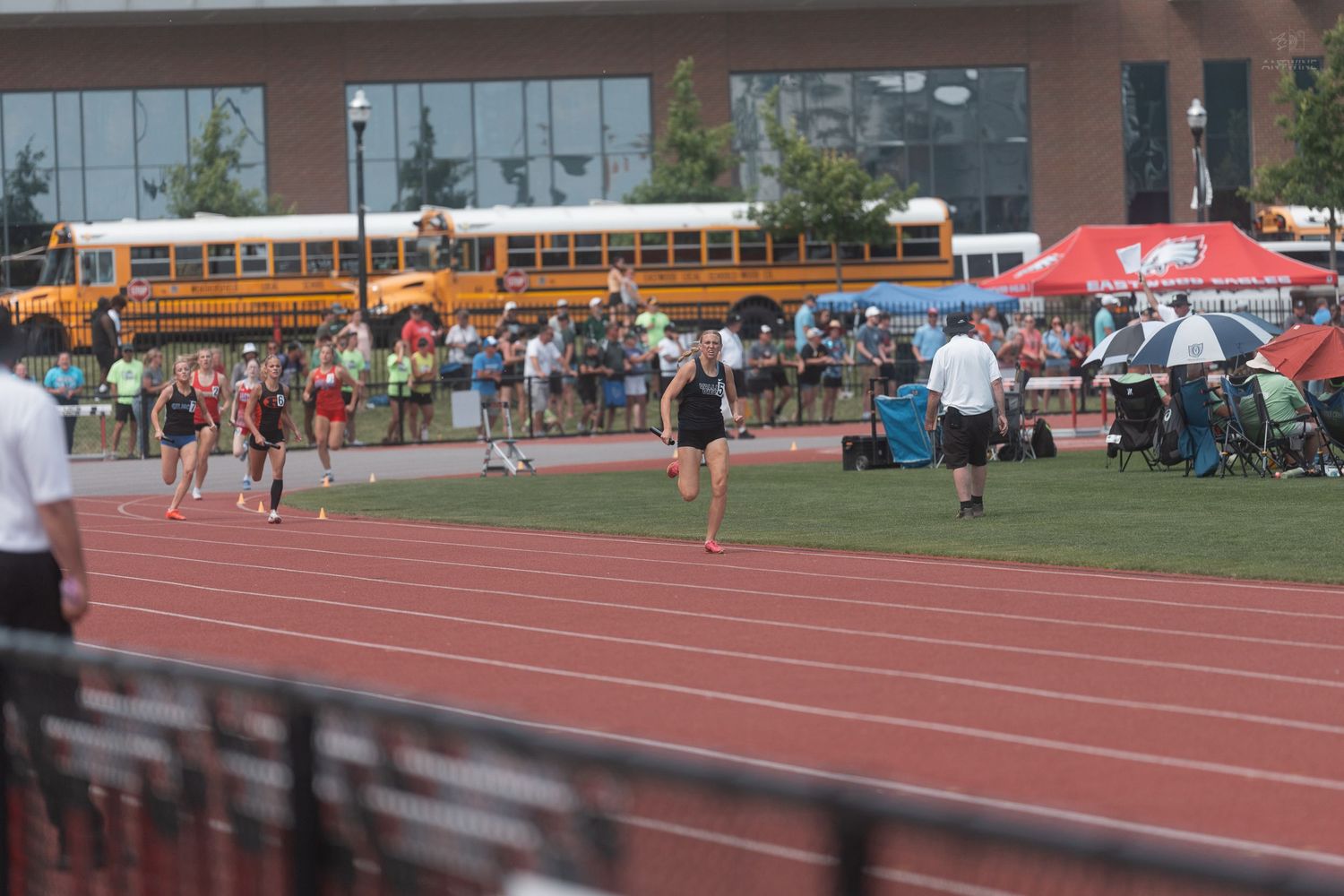 Athletes run on a red track at an outdoor meet with school buses and spectators visible in the background.