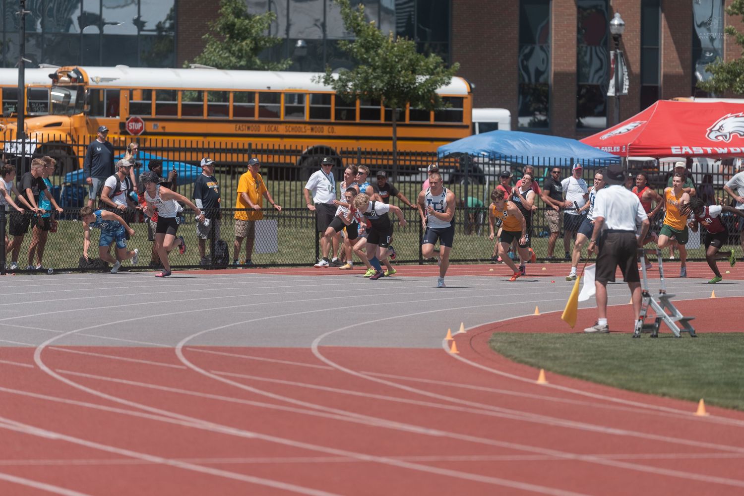 Young track athletes sprint past orange cones at the start of a race at an outdoor stadium meet.