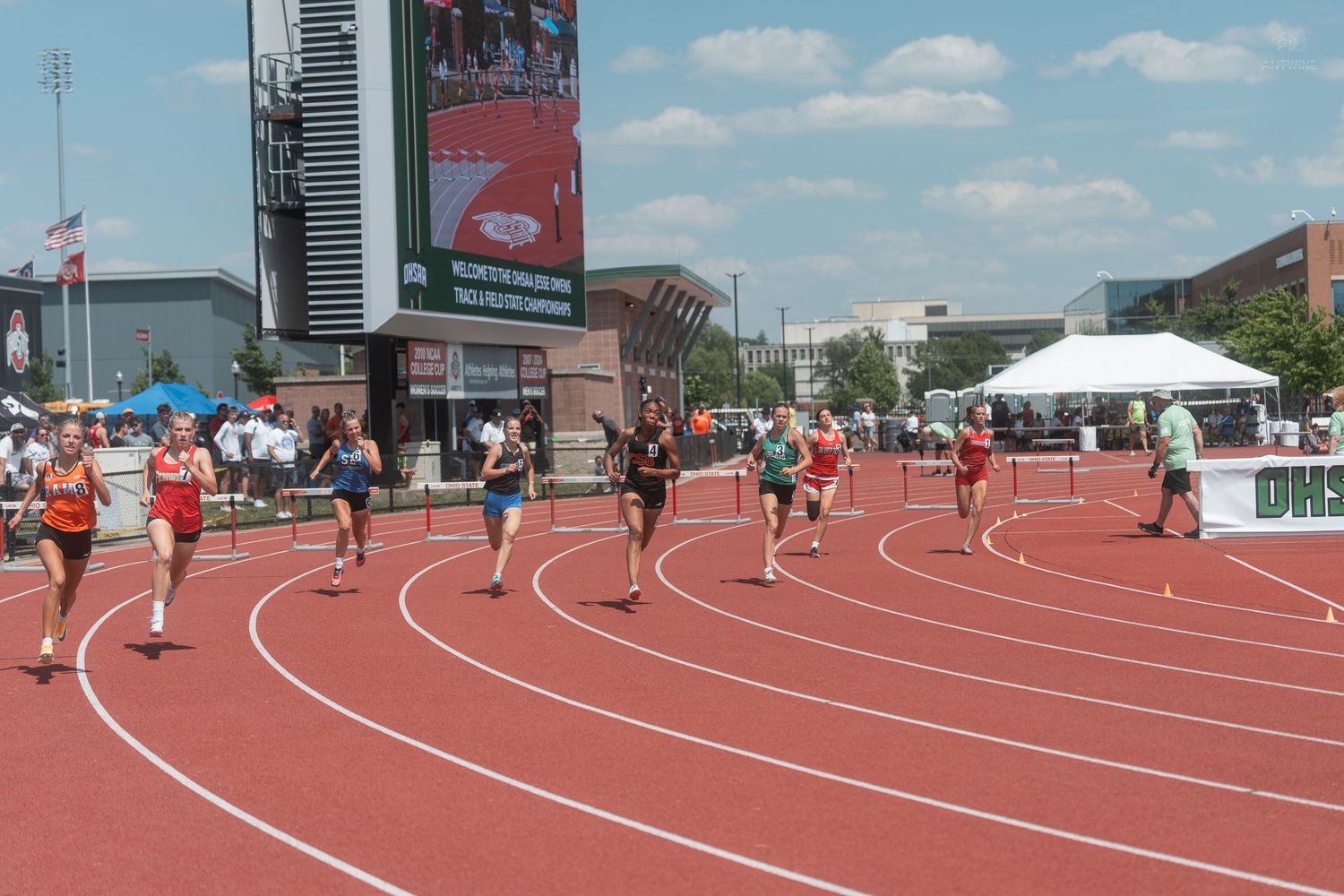 Multiple athletes competing in a track race on a red outdoor track with a large scoreboard visible in background.