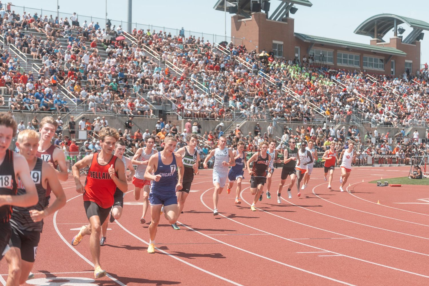 Large group of runners sprint down a red track at a high school track meet with packed stadium stands.