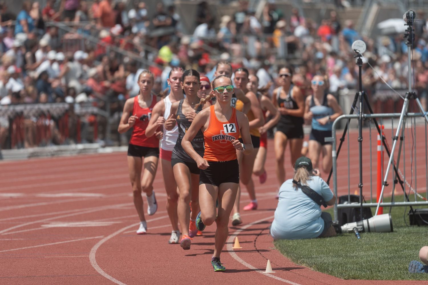 Female athletes race on a red track led by a runner in orange at a crowded outdoor track and field meet.
