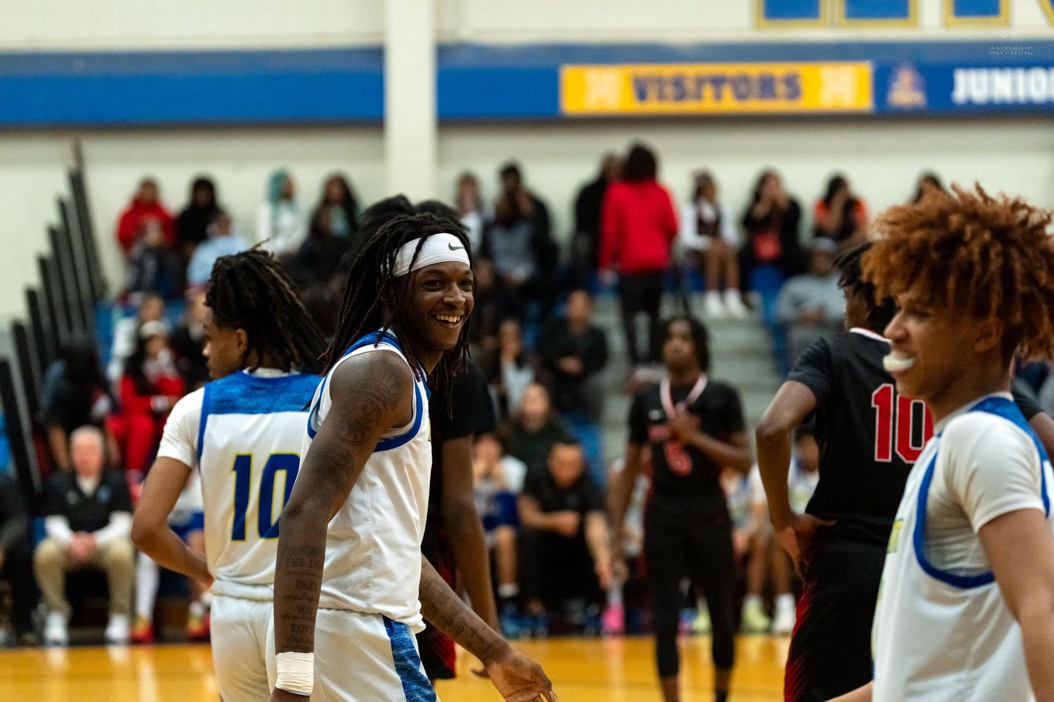 High school basketball players smile and interact on the court during a game with fans in the background.