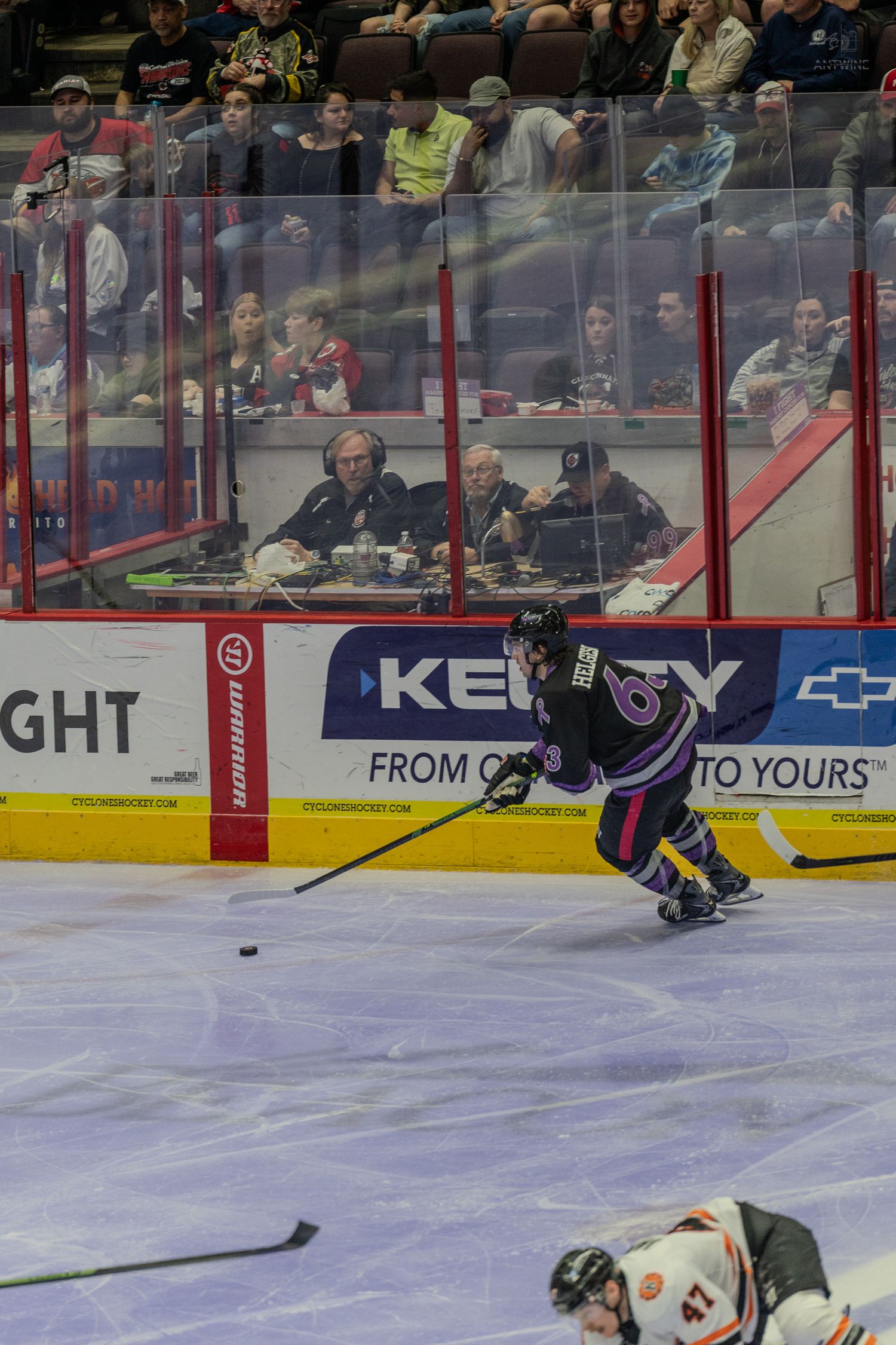 Hockey player in purple uniform skates with the puck near the boards with commentators visible behind the glass.