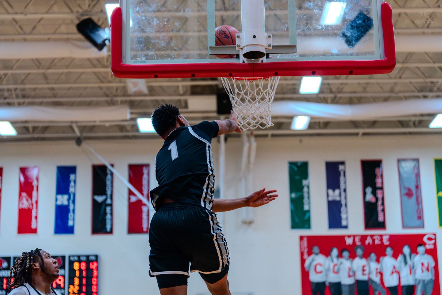 Basketball player in black uniform completes a dunk during a high school gymnasium basketball game.