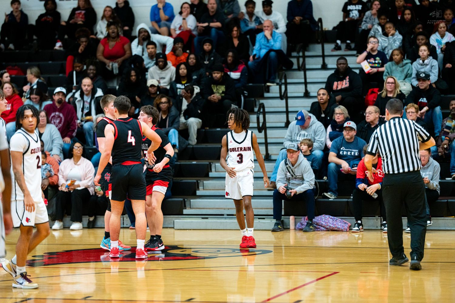 Basketball players and referee stand on court during a high school game with fans in bleachers.