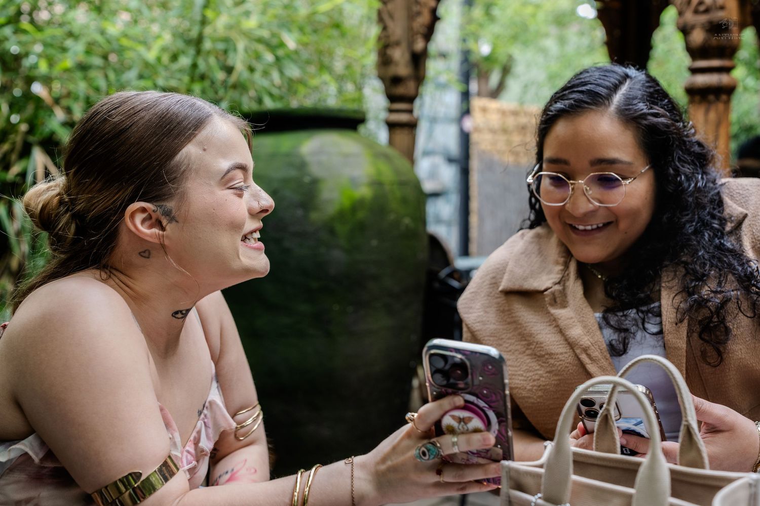 Two smiling women sharing phone screen at outdoor garden cafe table together.