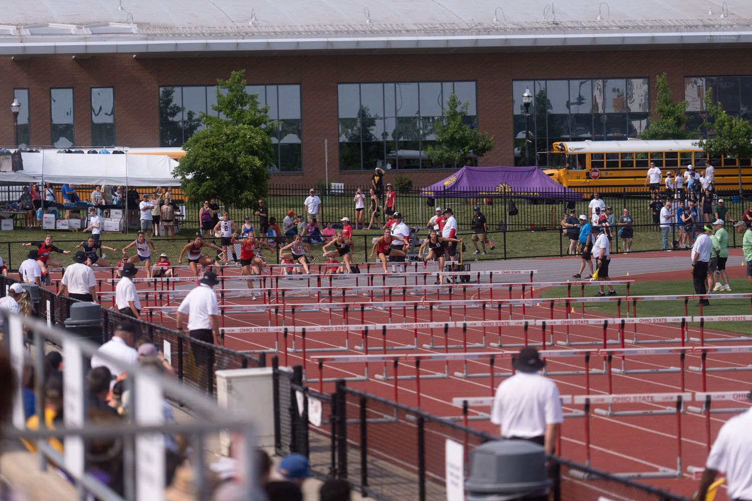 Wide shot of hurdles race in progress at outdoor track meet with spectators lining the field.