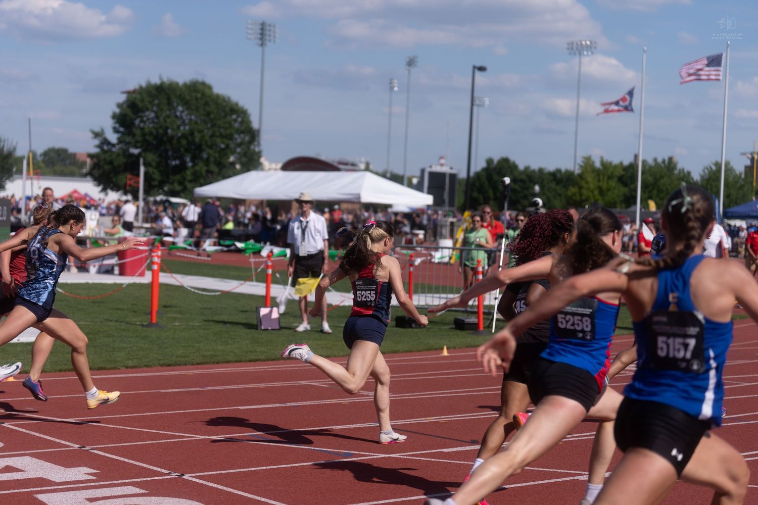Athletes sprint toward the finish line during a competitive outdoor track and field relay race on a sunny day.