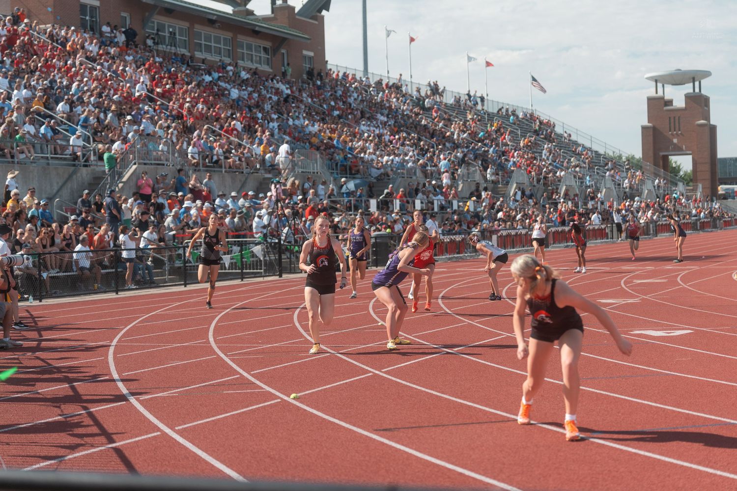 Runners compete in a relay race on a red track at a large outdoor stadium filled with spectators.