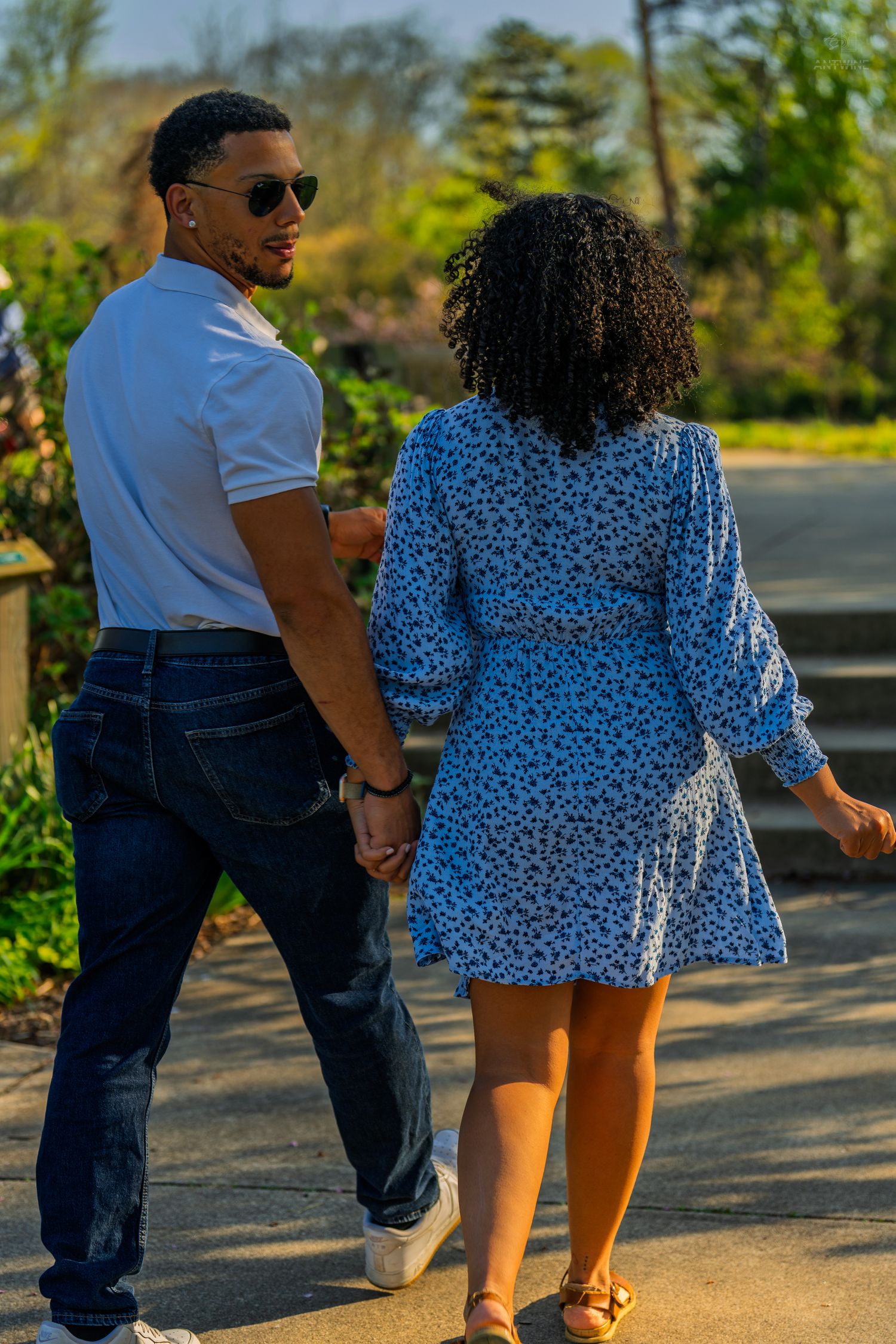 Man in sunglasses and white polo holds hands with a woman in a blue floral dress in a sunny park.