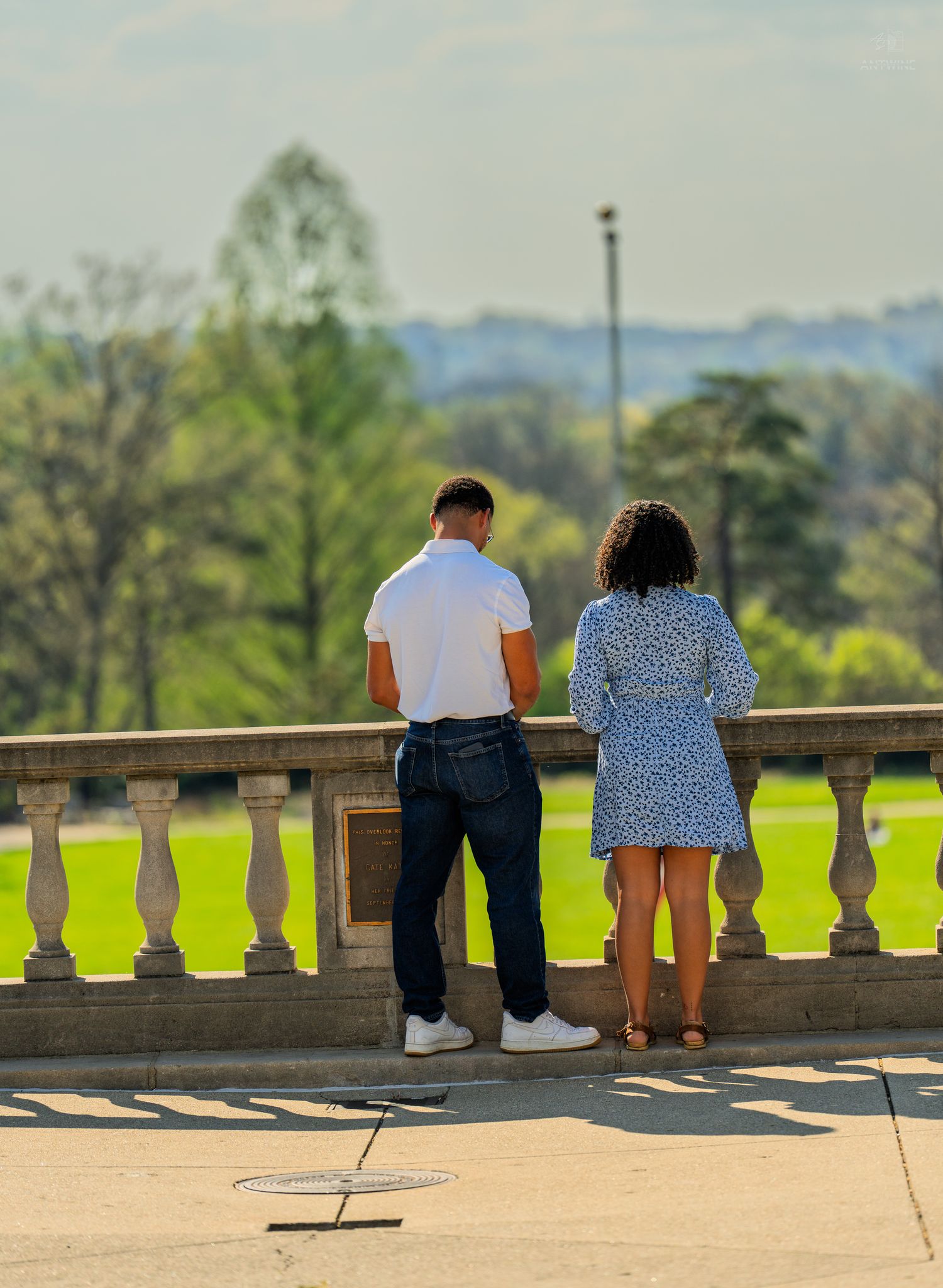 Man and woman in summer attire admiring green landscape from ornate stone terrace.