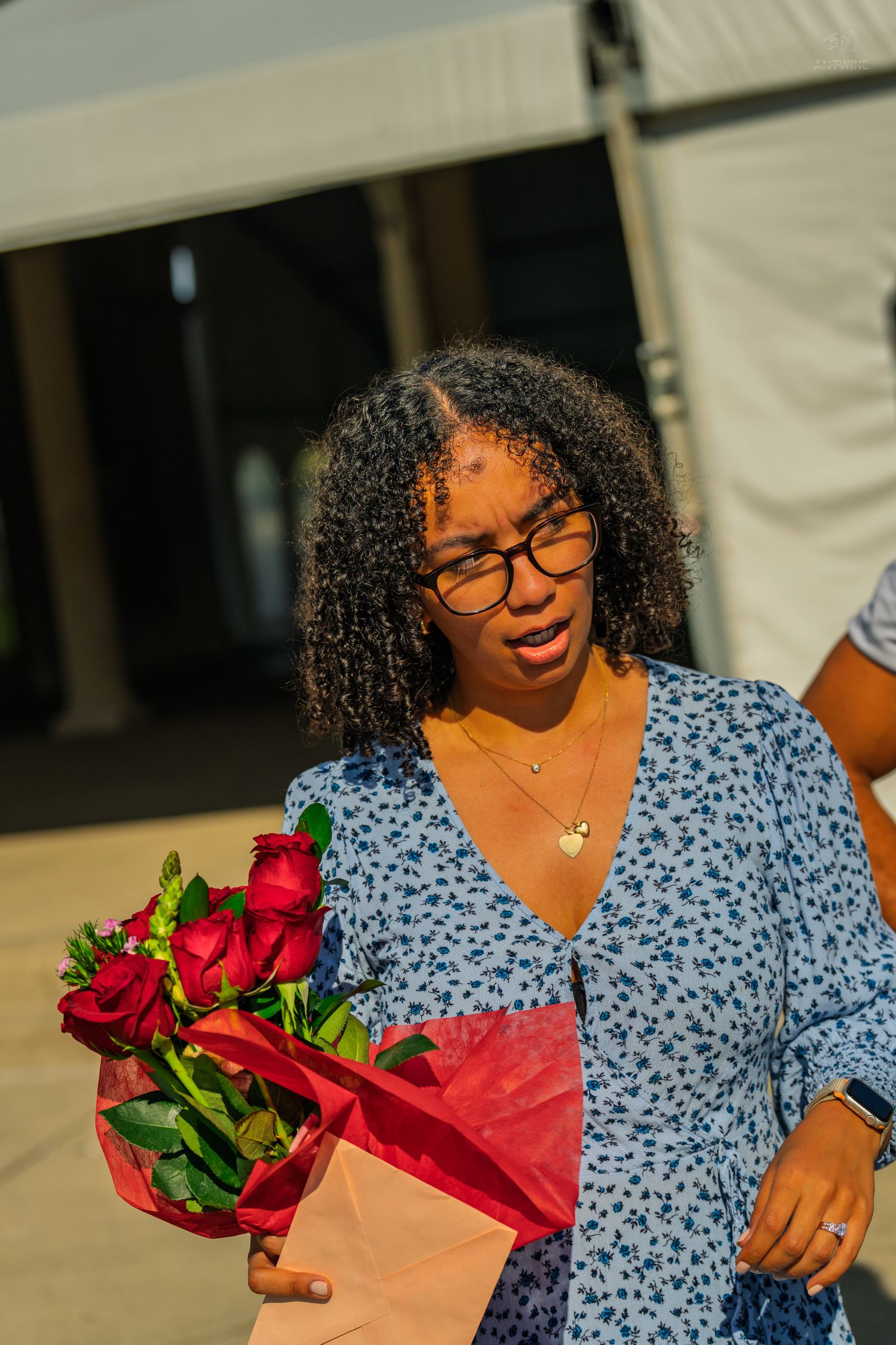 A young woman with curly hair and glasses holds a bouquet of red roses wrapped in red paper outdoors.