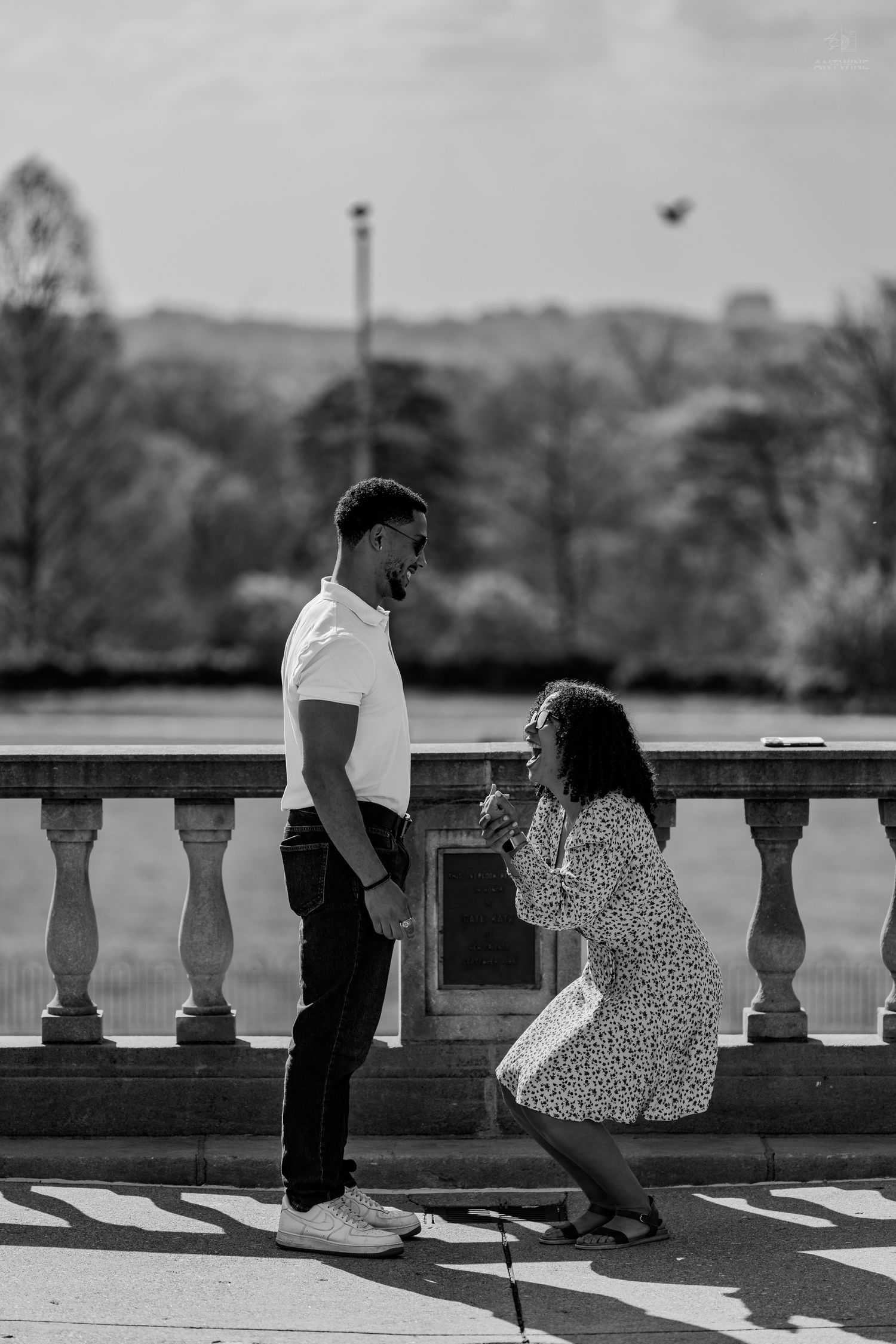 Black and white photo of woman kneeling before man holding ring box in outdoor park proposal.