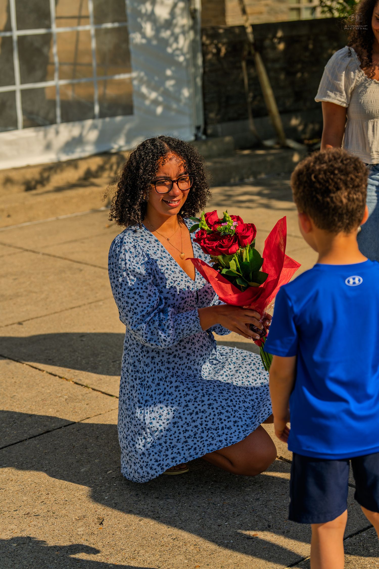 Smiling woman in blue floral dress kneels to receive red roses from a young boy in blue shirt outdoors.